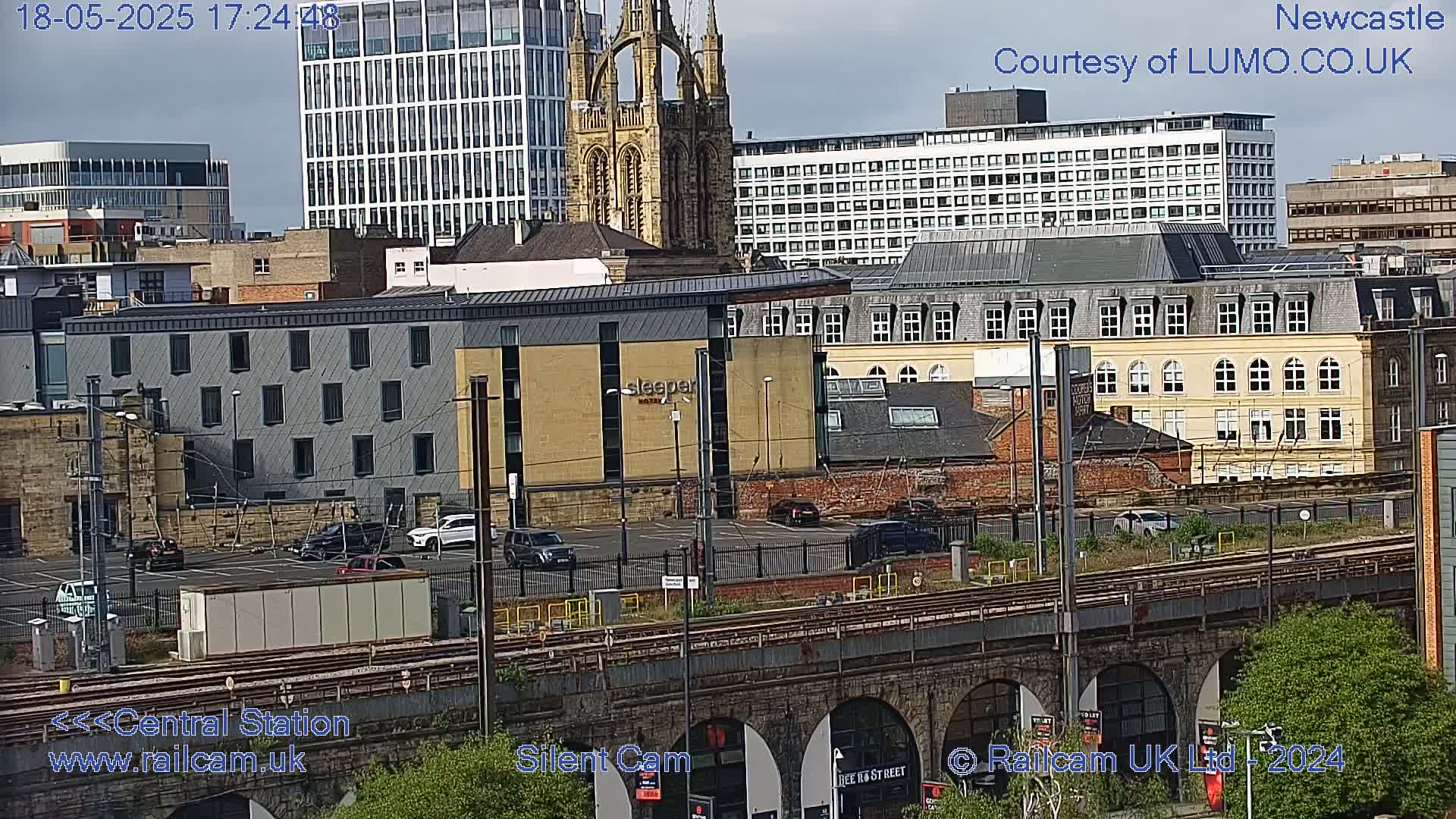 A city view on an overcast day shows a mix of modern and older buildings surrounding a railway track and bridge.