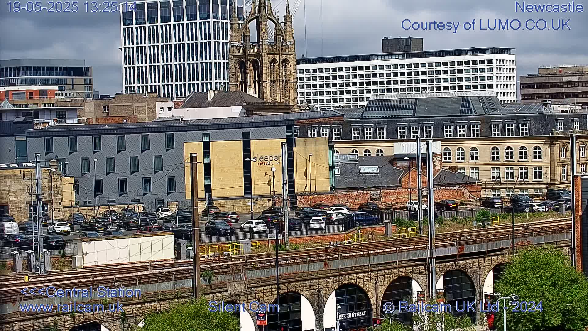 A city scene on a partly cloudy day shows various buildings, including a high-rise, a church, and a hotel, with cars parked in a lot near a railway viaduct.