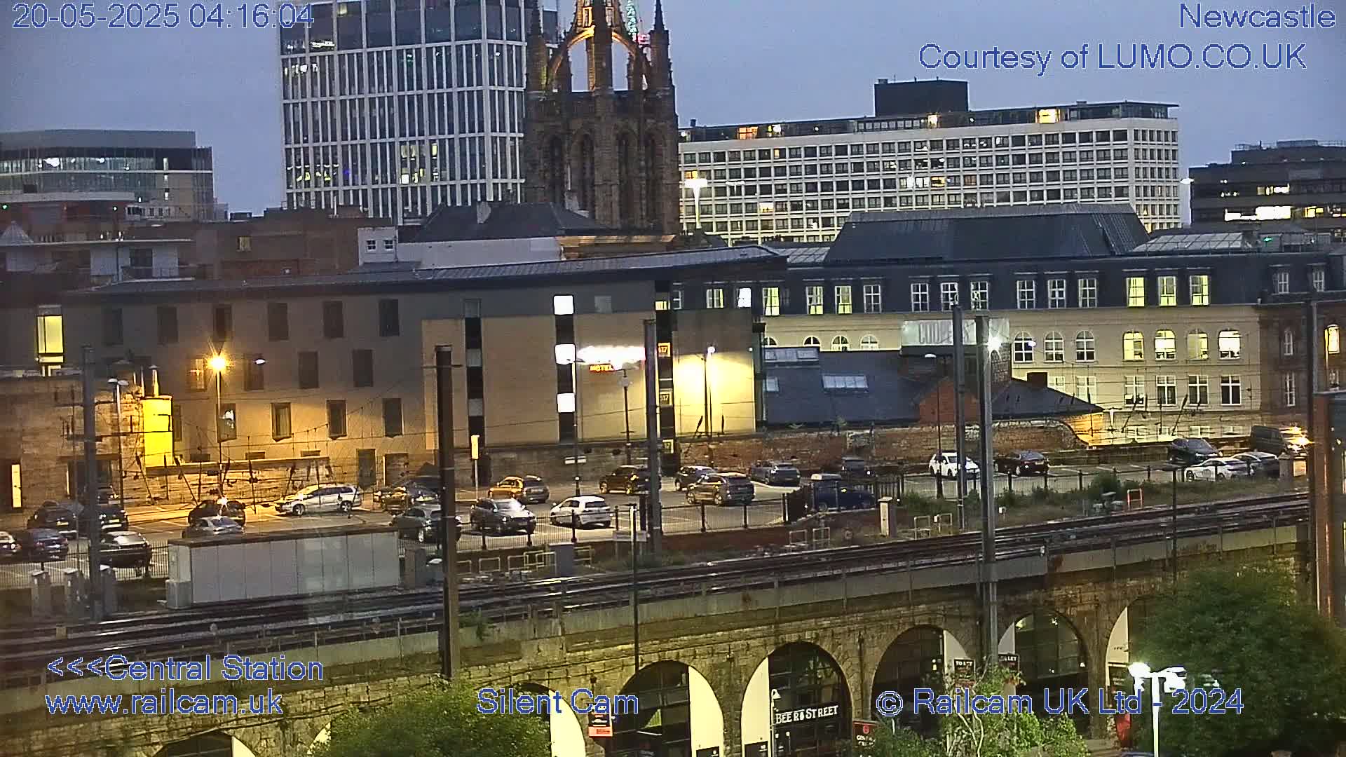 A nighttime cityscape with illuminated buildings, cars parked in a lot, and a railway track with arched bridge, under a dusky sky.