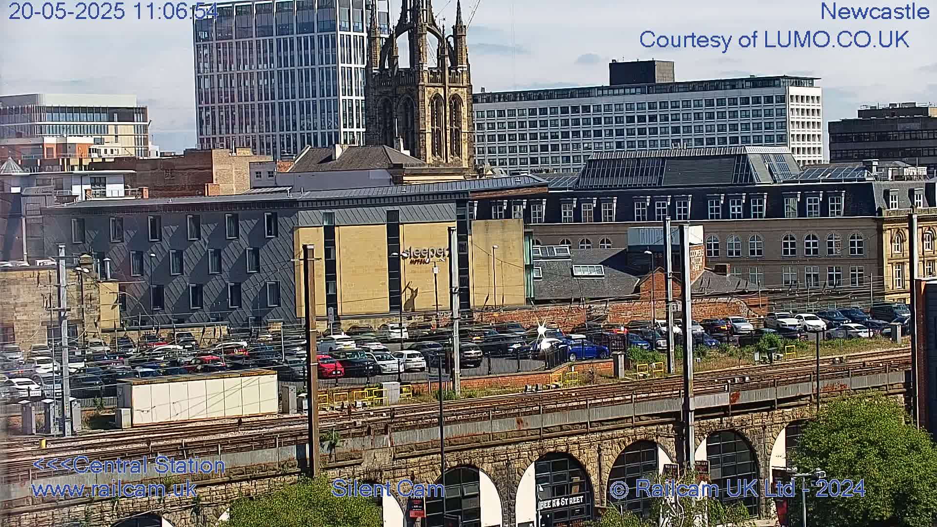 A city scene on a sunny day, showing a large parking lot filled with cars next to a railway viaduct and various buildings including a church spire.
