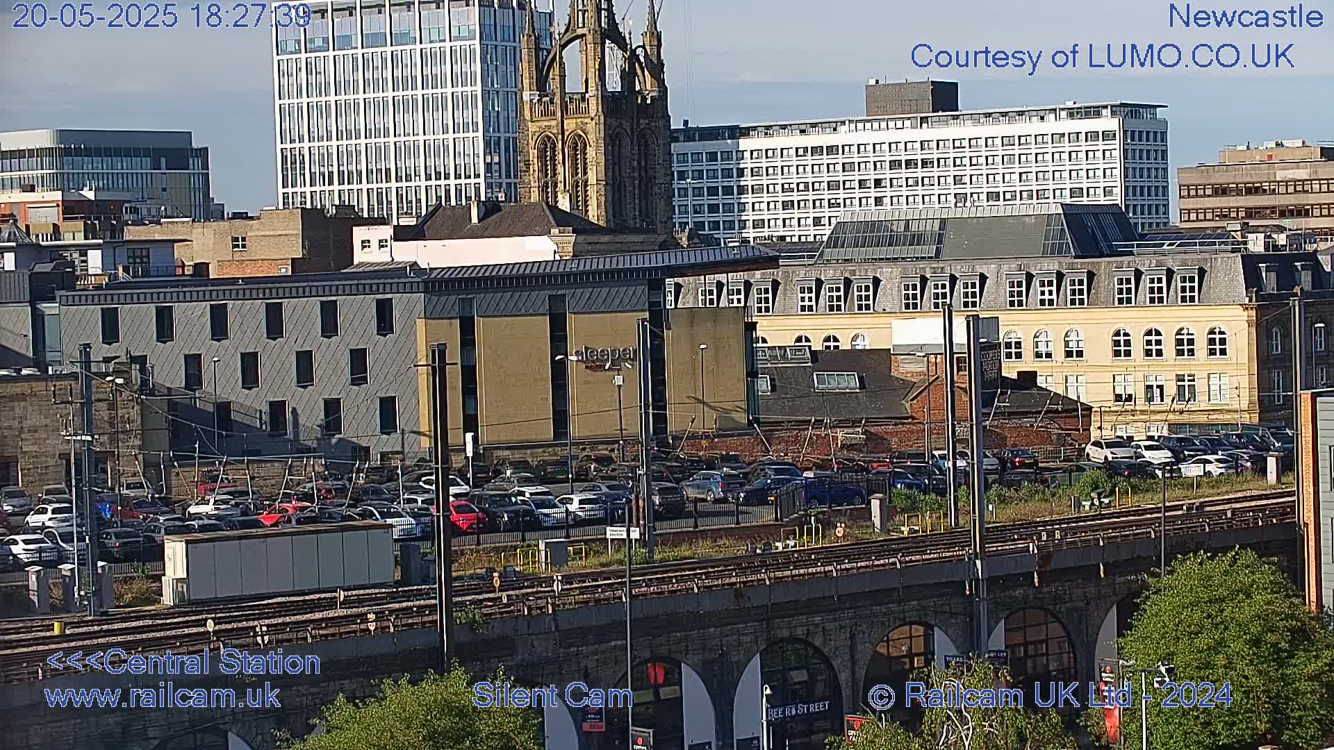 A cityscape of Newcastle, under a mostly clear sky, shows a train station, railway tracks, various buildings, and a large parking lot filled with cars.