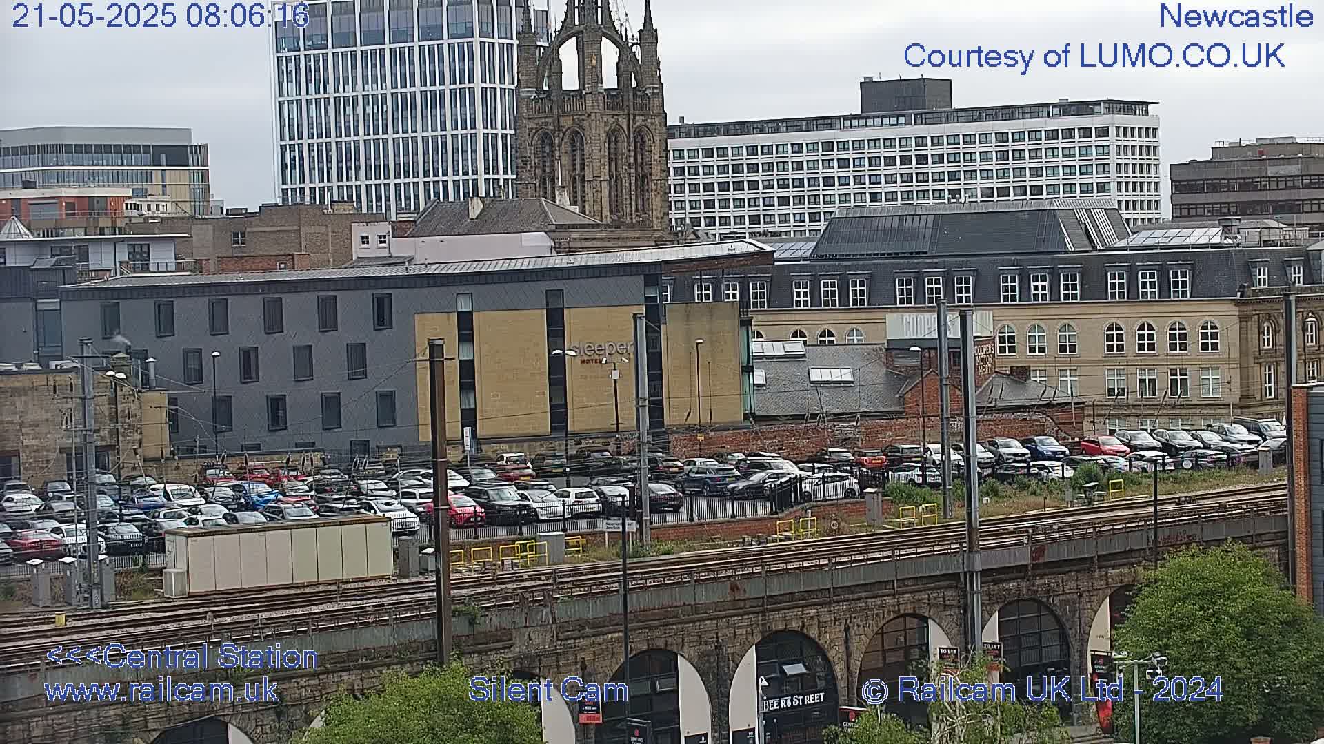 A city landscape viewed from above shows a large parking lot filled with cars next to train tracks and a bridge, with various buildings, including a tall modern building and a stone church, in the background under an overcast sky.