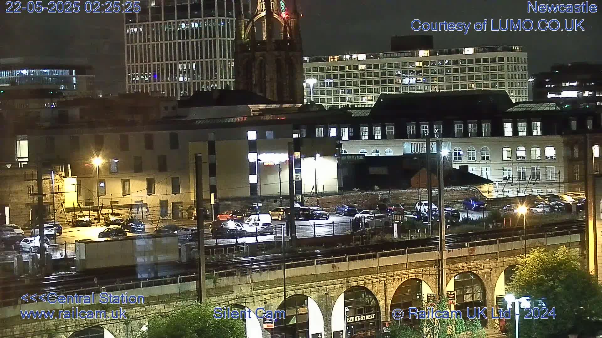 A nighttime view of a city's central station area, showing a parking lot with several cars, train tracks below a stone arch bridge, and various buildings in the background under a clear sky.