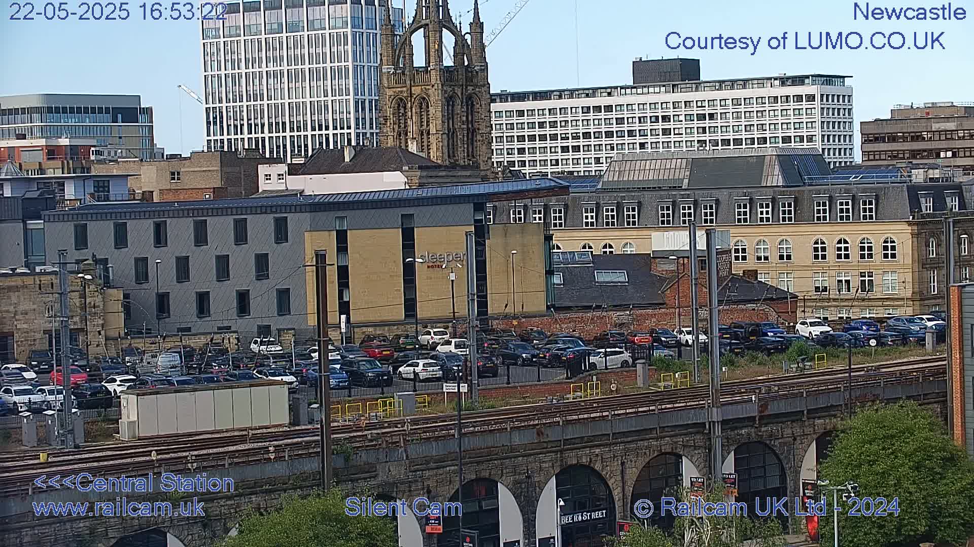 A city scene on a partly sunny day shows a large parking lot filled with cars next to train tracks, with various buildings including a tall church in the background.