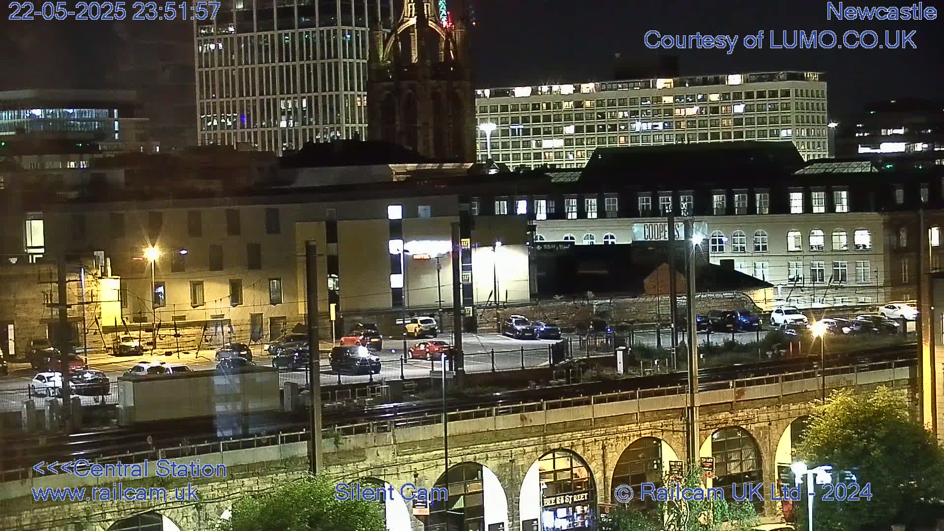 A nighttime view of a city's cityscape, featuring buildings, parked cars, and a railway viaduct.