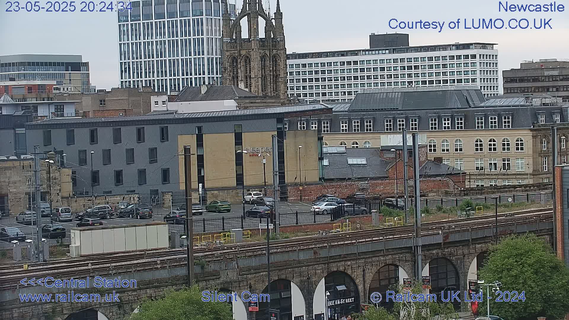 A view of a city with overcast skies, showing a mix of modern and older buildings, a parking lot with several cars, and train tracks on a viaduct.