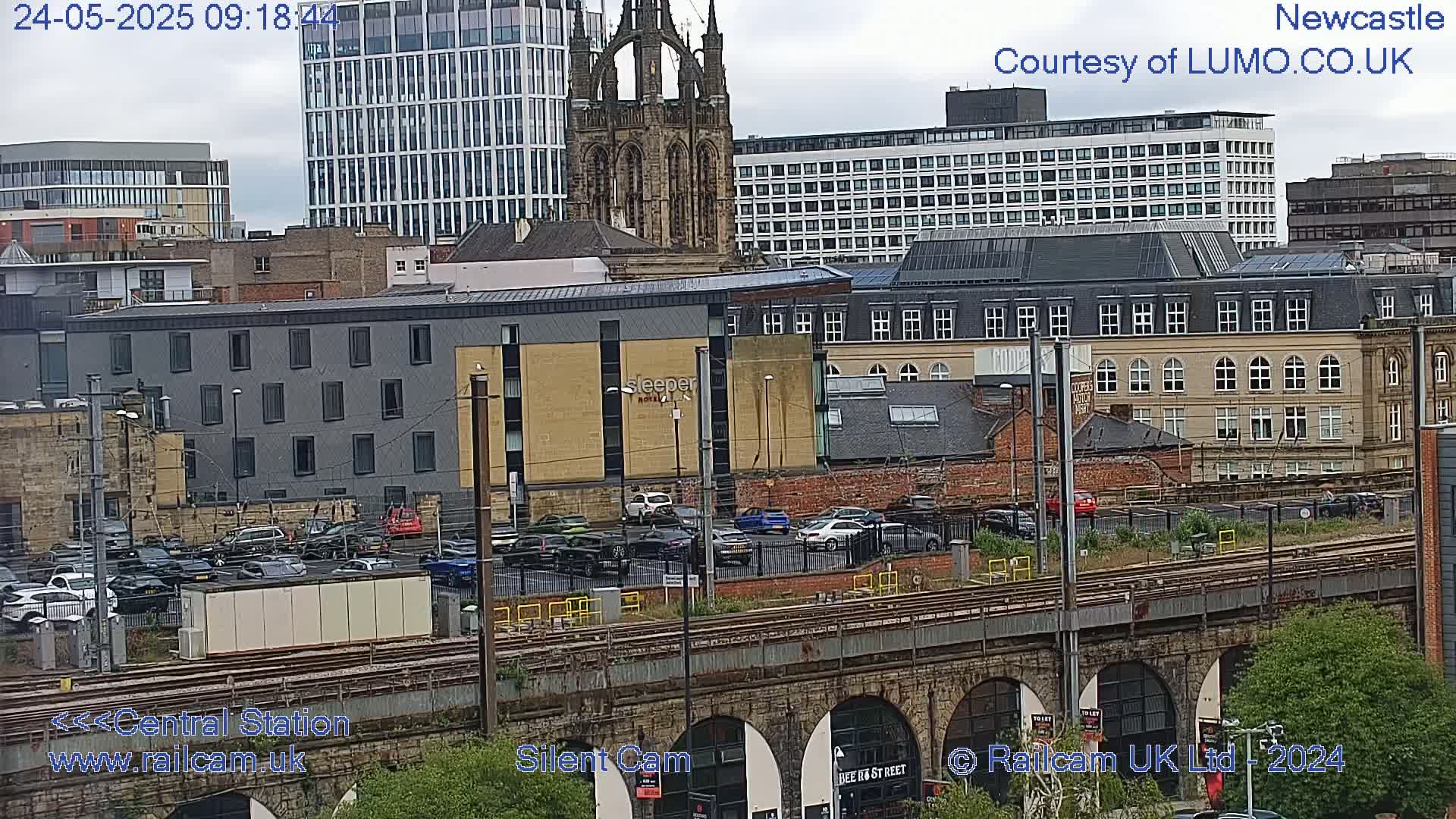 A city scene viewed from above shows a parking lot, train tracks, and a variety of buildings under an overcast sky.