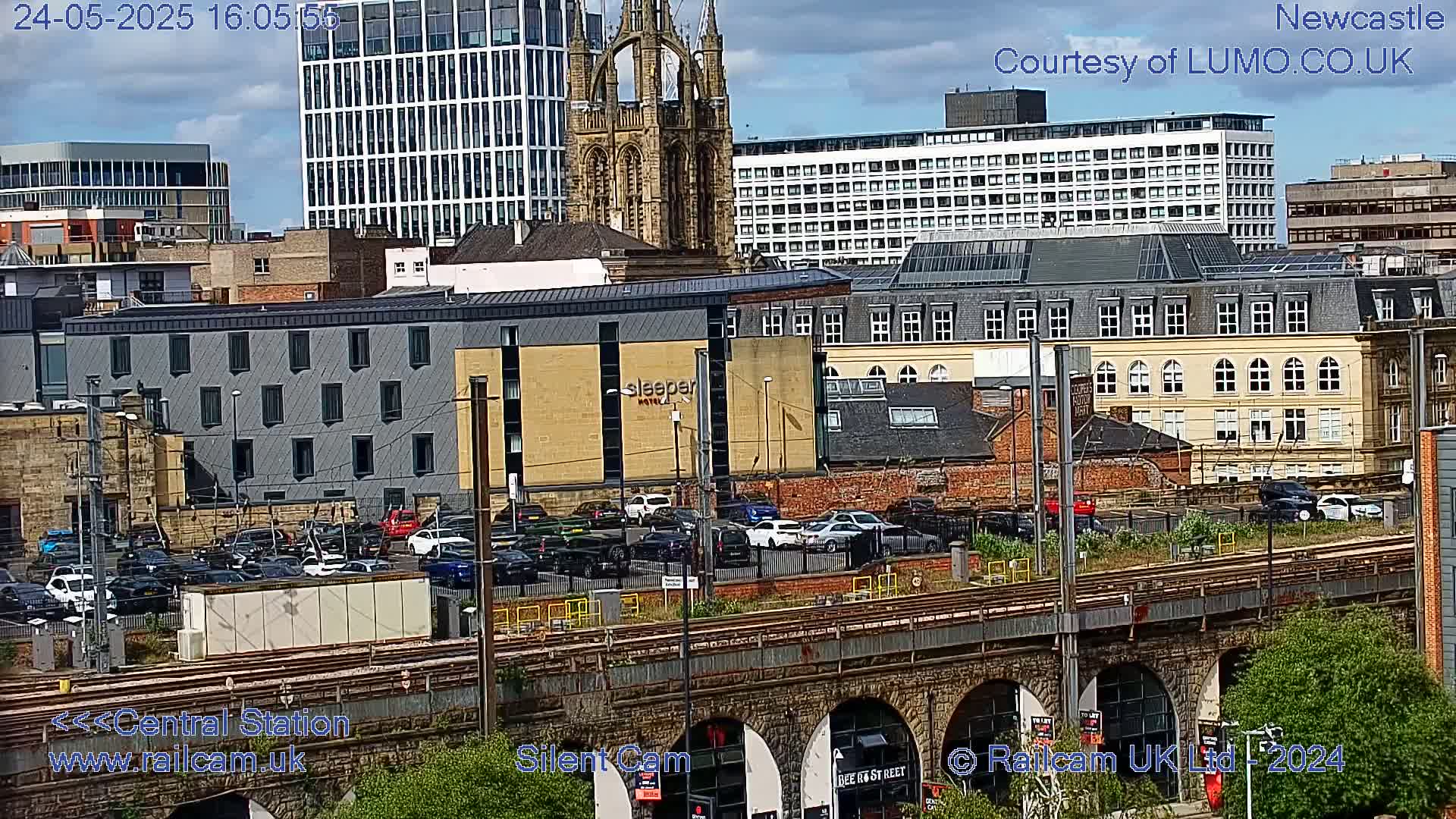 A city scene under partly cloudy skies shows a railway viaduct above a parking lot and various buildings, including a hotel and a cathedral.