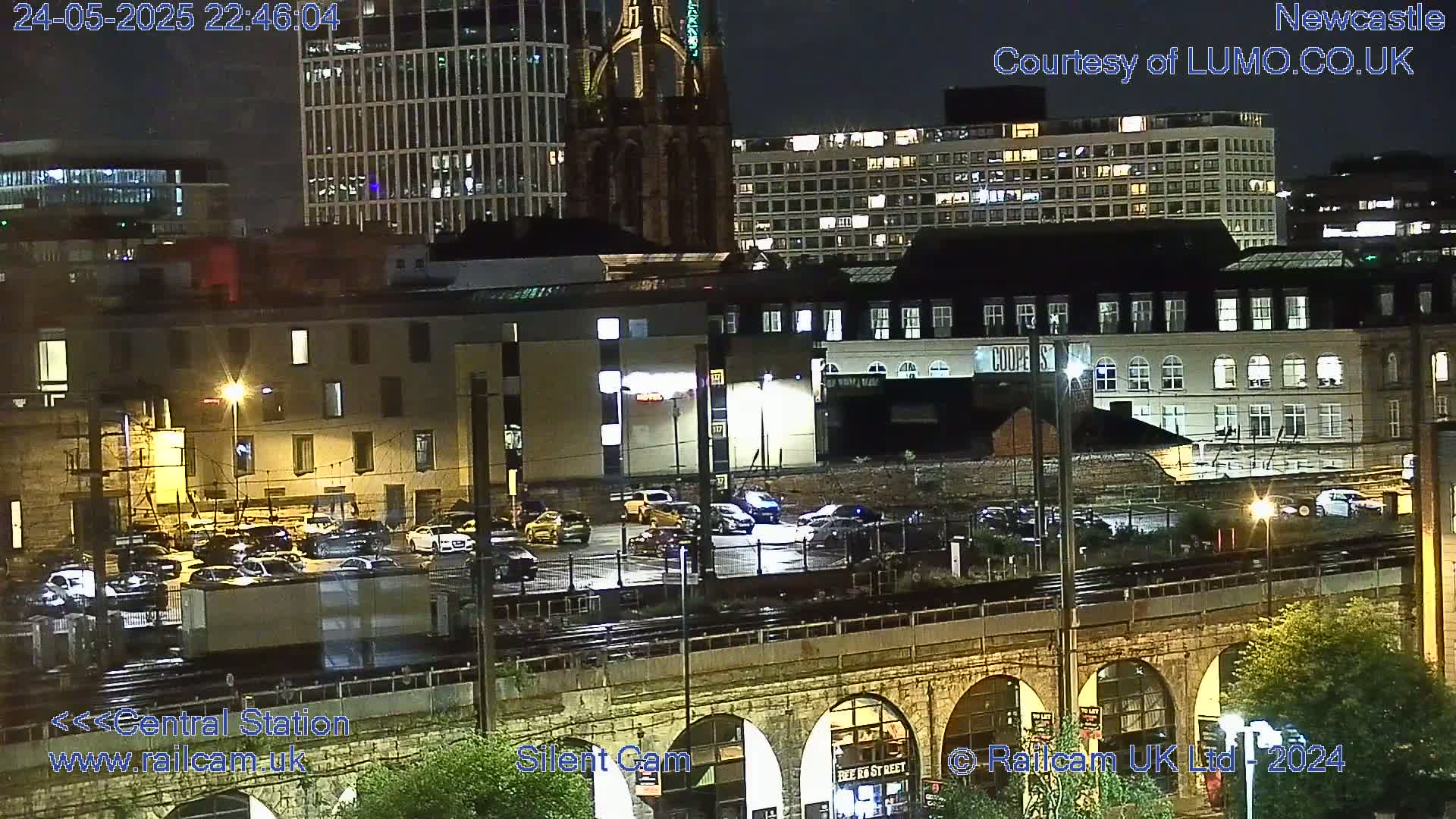 A nighttime view of a city’s illuminated buildings and a parking lot near train tracks, under slightly wet conditions.