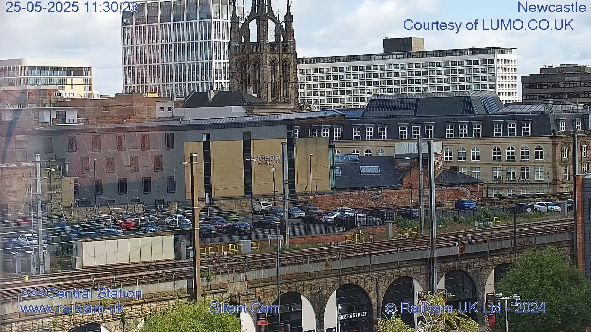 A city landscape under partly cloudy skies, showing a mix of modern and older buildings, a railway viaduct, and a parking lot filled with cars.