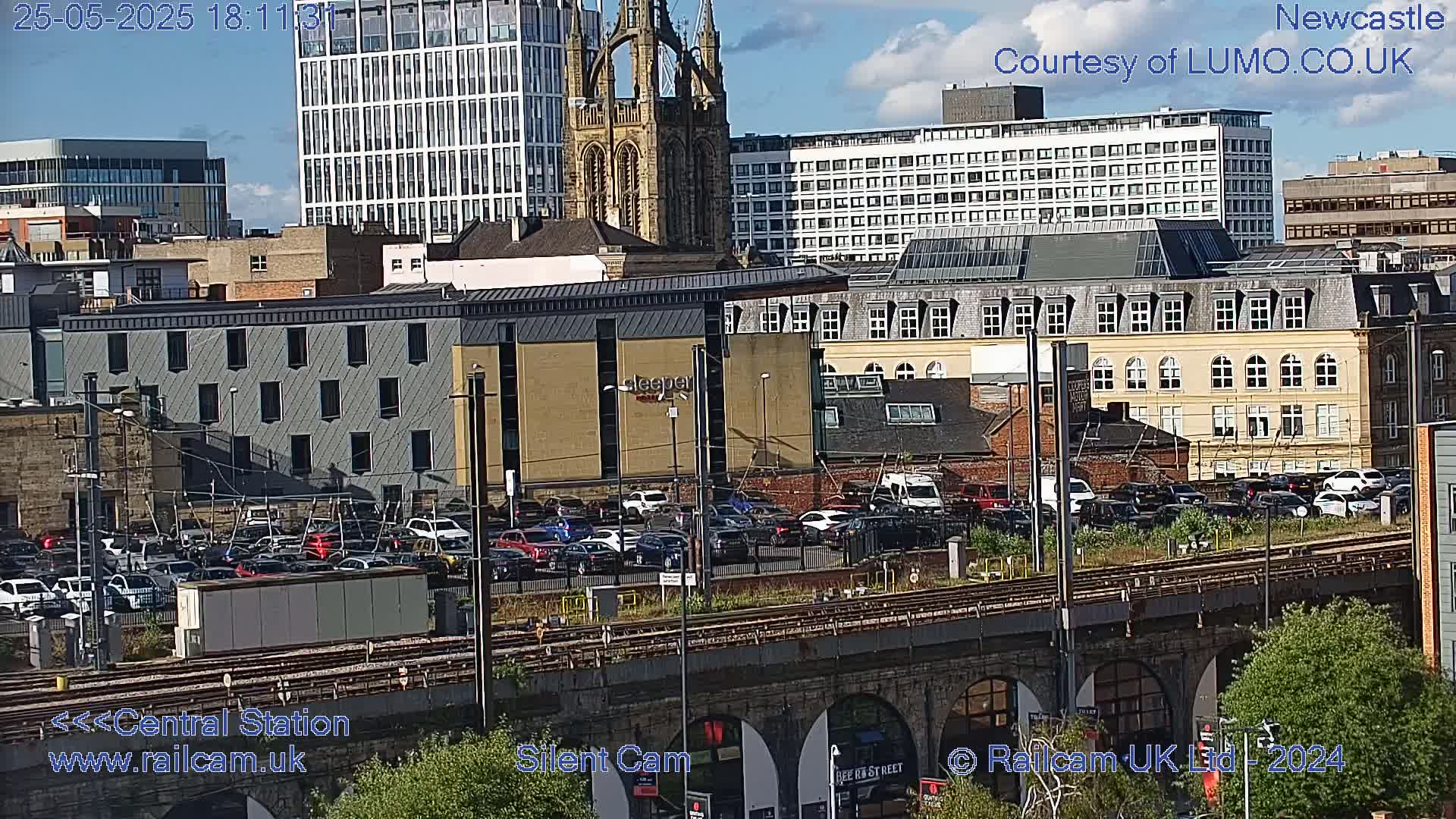 A partly cloudy daytime view of a city showing a large parking lot filled with cars, train tracks, and various buildings, including a tall modern building and a church.