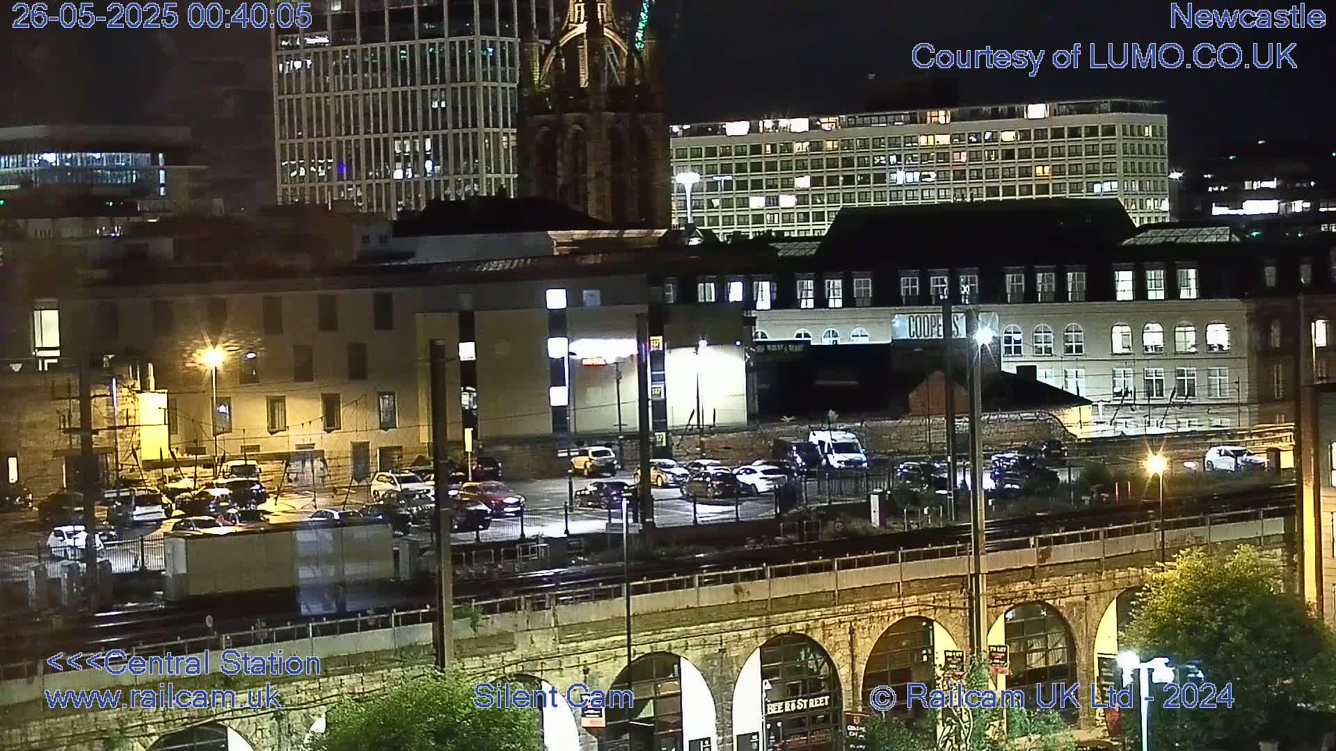 A nighttime view of a city's train station and surrounding buildings, with cars parked in a lot below a stone viaduct.