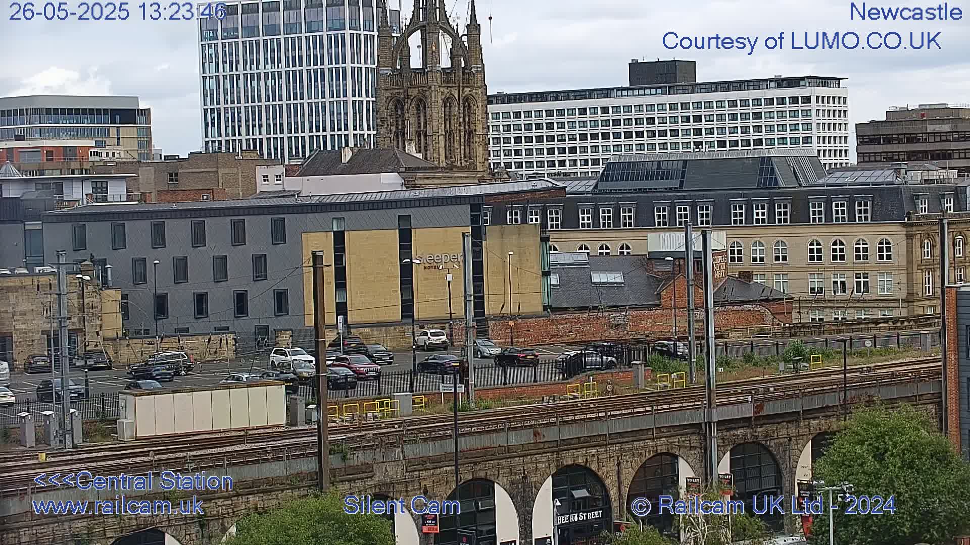 A city scene viewed from above, showing a mix of modern and older buildings, a railway line, and a parking lot under a partly cloudy sky.