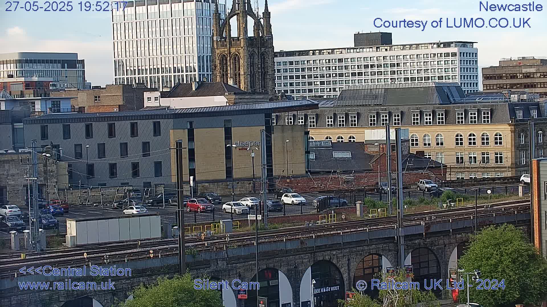 A city landscape under a partly cloudy sky, featuring a train track that passes under an arched bridge, a parking lot filled with cars, and several multi-story buildings including a church.