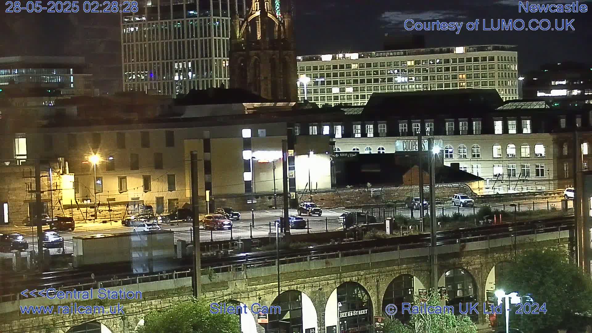 A nighttime view of a city featuring buildings, a parking lot with cars, and a train track elevated on a stone viaduct under a mostly clear sky.