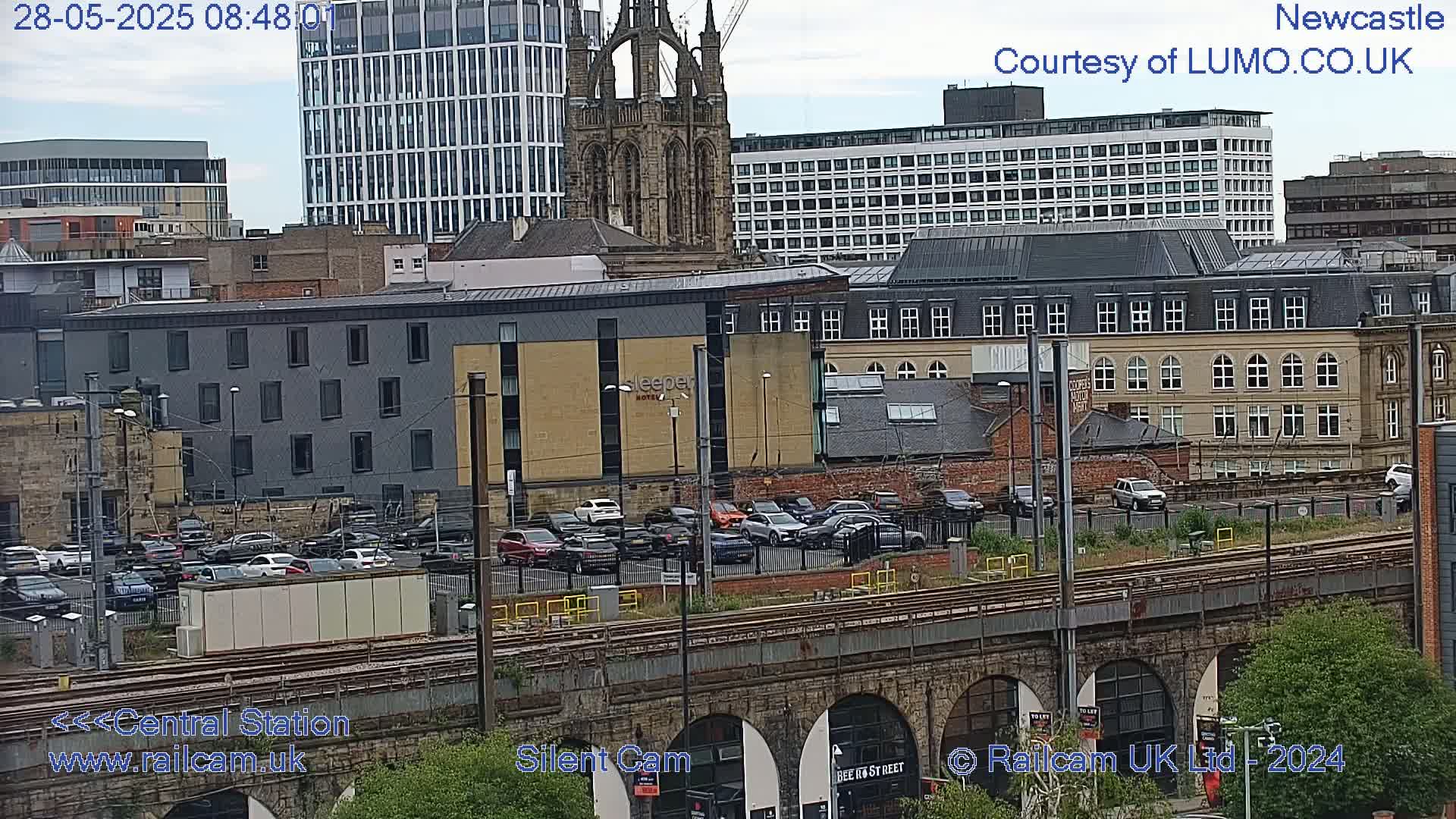 A city scene with a mix of modern and older buildings, a parking lot full of cars, train tracks, and stone arches, under a partly cloudy sky.