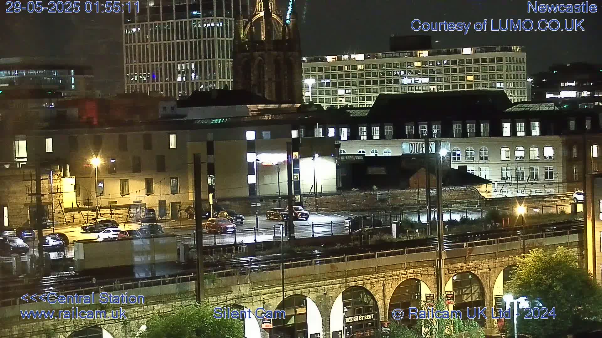 A nighttime view of city buildings and a parking lot, illuminated by streetlights, with a train track and stone archway in the foreground.