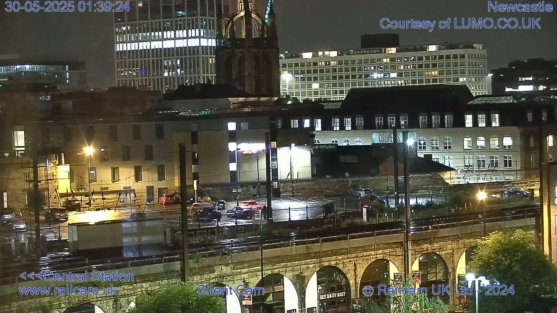 A nighttime view of a city featuring several buildings, a railway, and a stone arch bridge under rainy conditions.