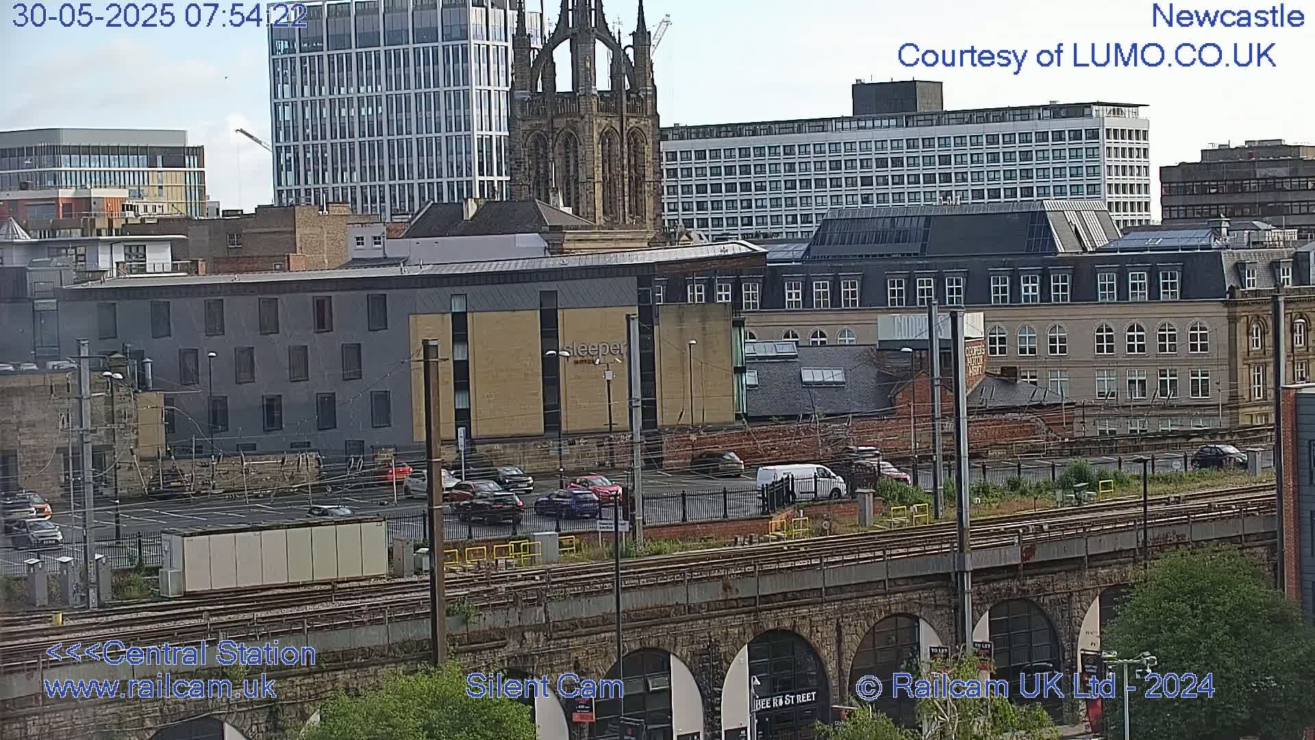 A city view on a partly cloudy day shows several buildings, including a tall modern structure, an older Gothic-style church, and a hotel, with a railway line and a parking lot in the foreground.