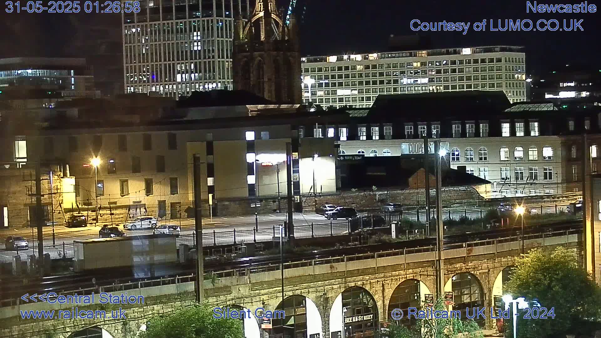 A nighttime view of a city's cityscape, showing buildings, a parking lot, train tracks, and a stone archway bridge.