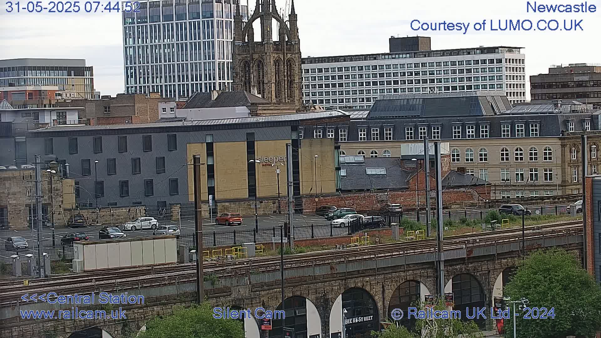 A city scene viewed from above, showing buildings, a parking lot with cars, train tracks, and a stone arch bridge under an overcast sky.