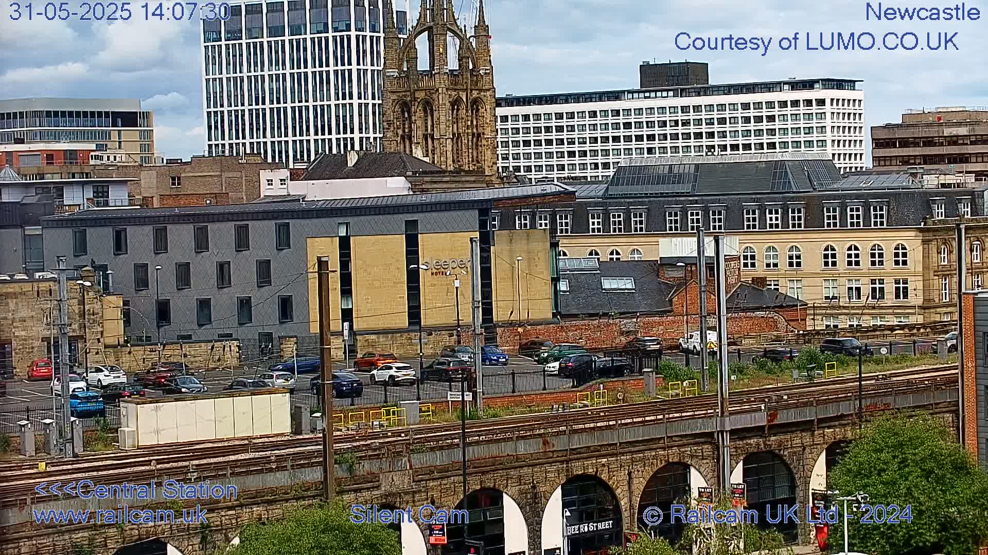 A city view on an overcast day shows a parking lot, train tracks, and various buildings including a modern hotel and a stone cathedral.