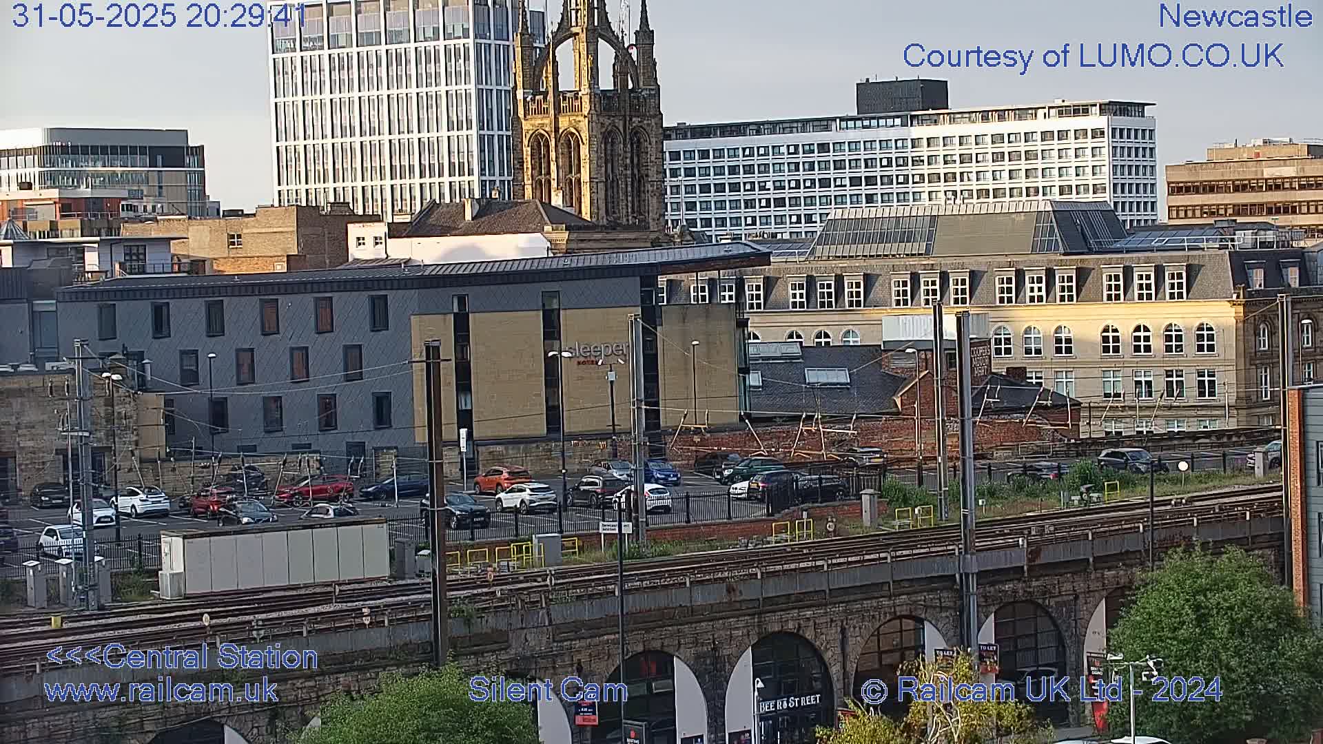 A city skyline at dusk, showing various buildings including a tall modern structure and a church, with a parking lot and train tracks in the foreground.