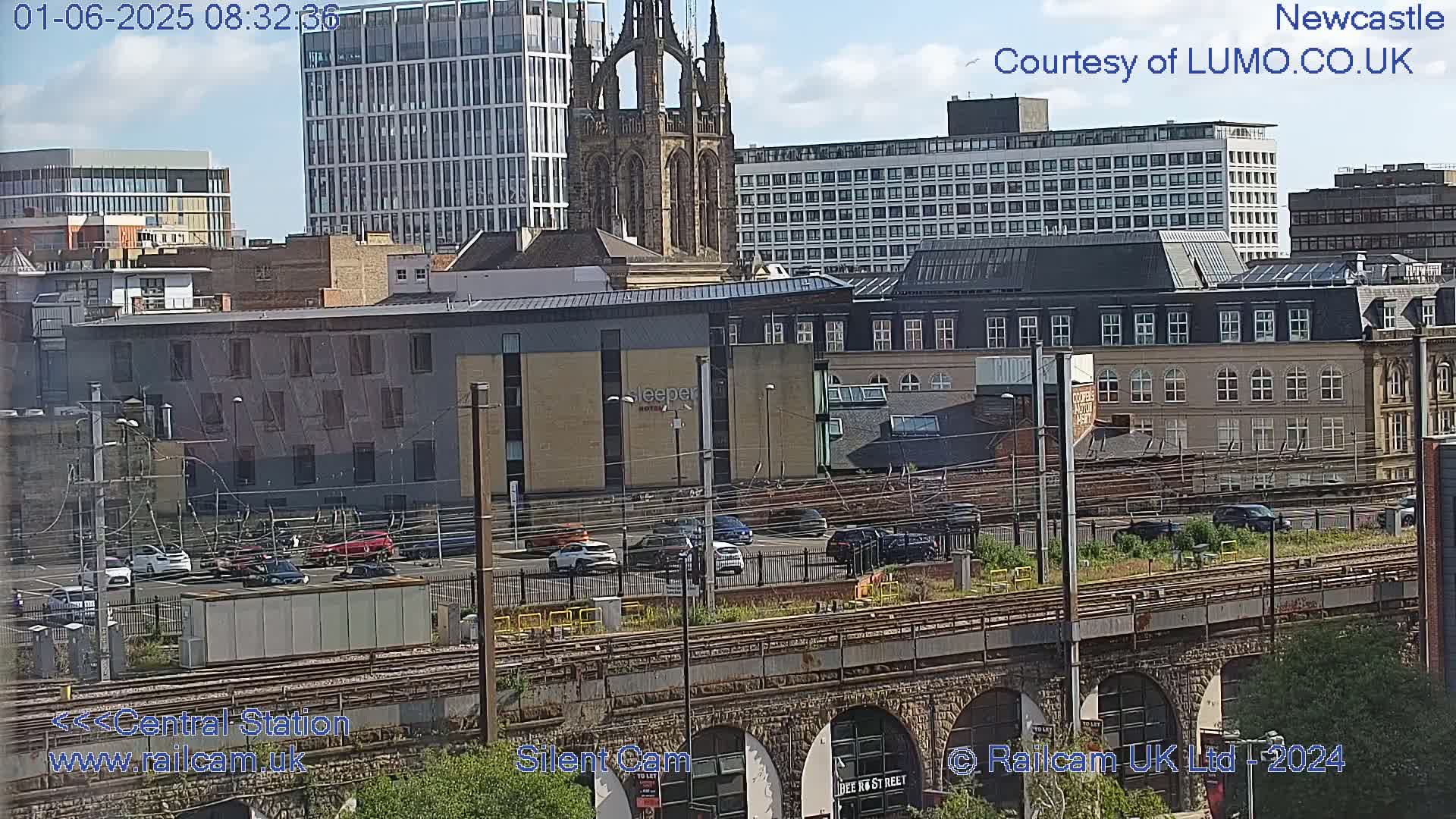 A city landscape under a partly cloudy sky shows a mix of modern and older buildings, railway tracks, and a parking lot with several cars.