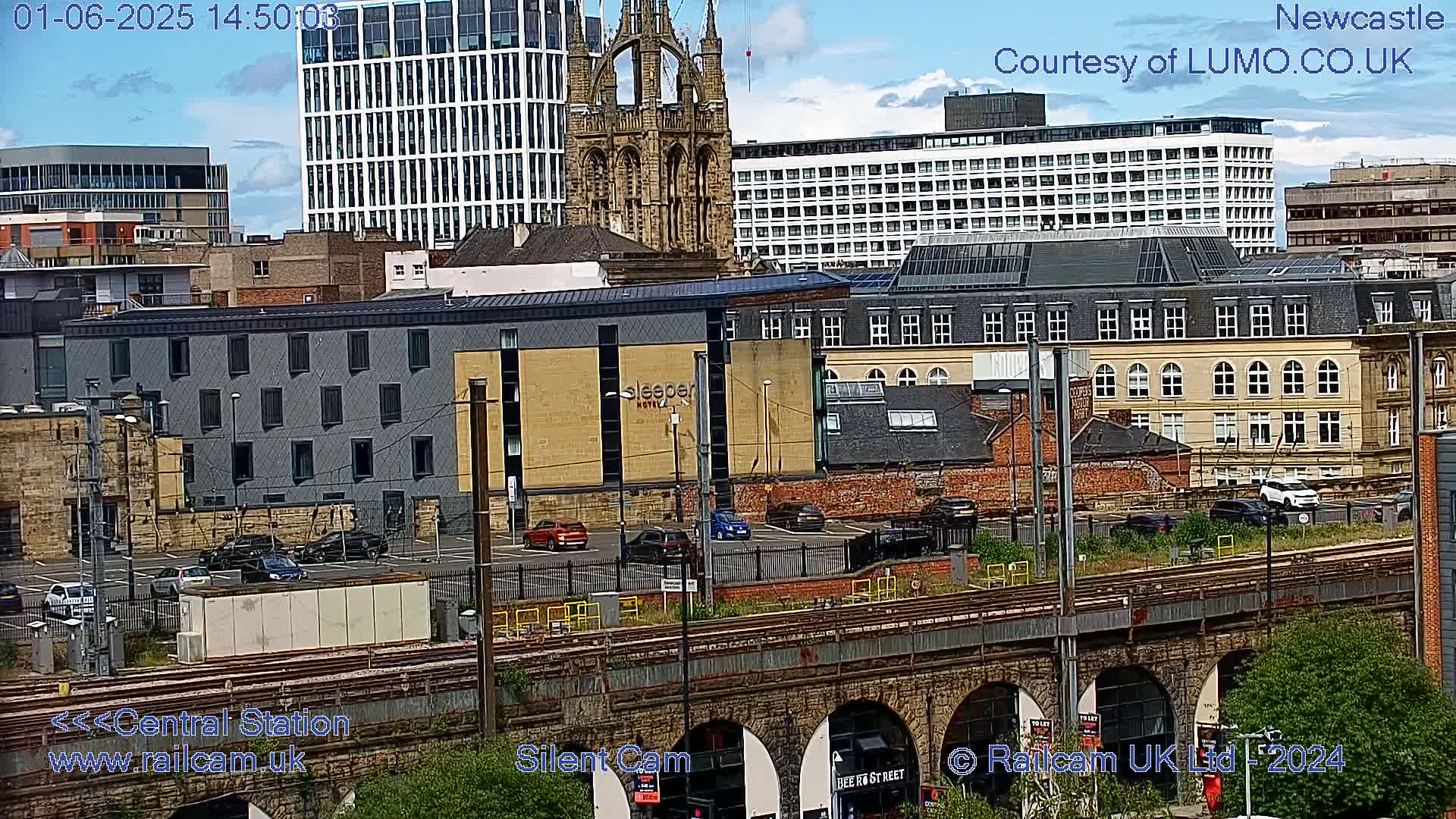 A city scene on a partly cloudy day, showing various buildings including a large church, modern high-rises, and older stone structures, with a railway line and viaduct in the foreground.