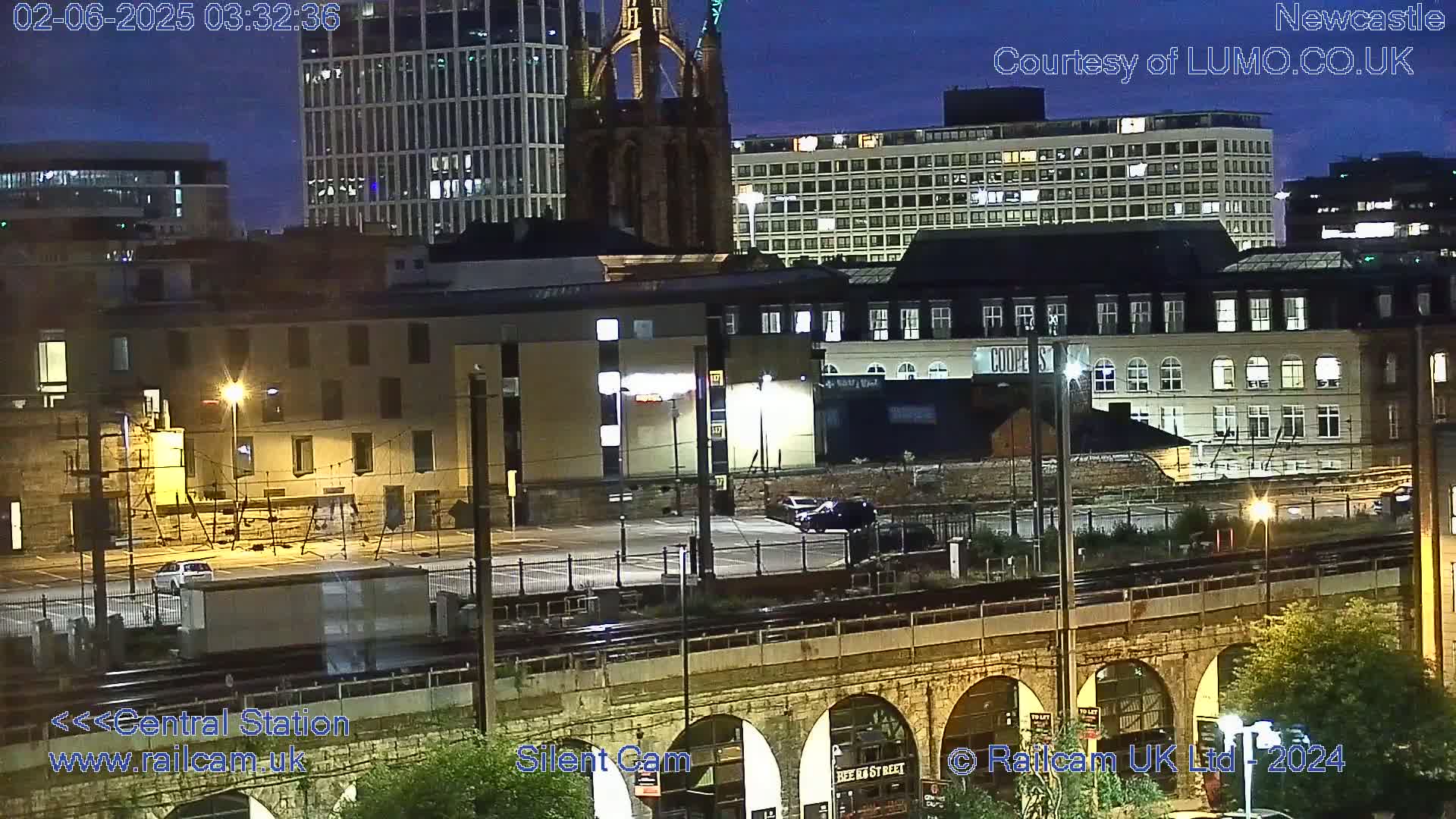 A nighttime view of a city with several buildings, a parking lot, train tracks, and a stone bridge under a dark blue sky.