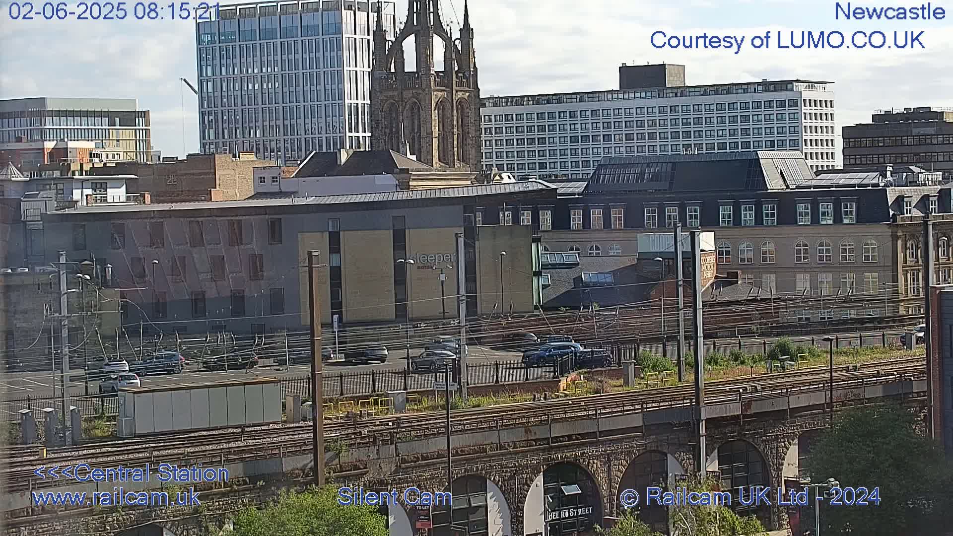 A city skyline under a partly sunny sky, featuring several multi-story buildings, a church, and train tracks with a bridge.