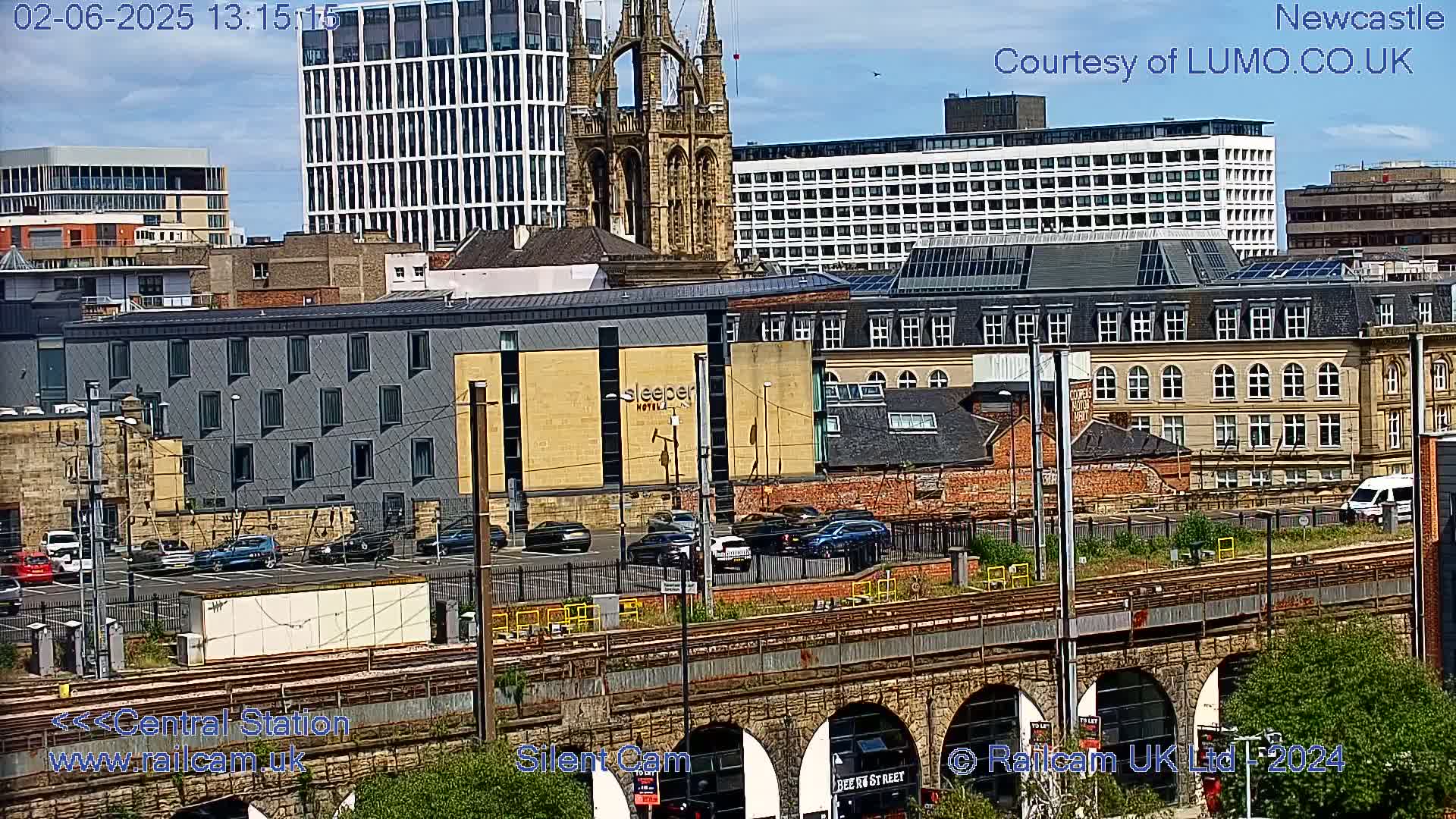 A city scene on a partly sunny day shows several buildings, including a large church and a hotel, with a train track and viaduct in the foreground and several cars parked nearby.