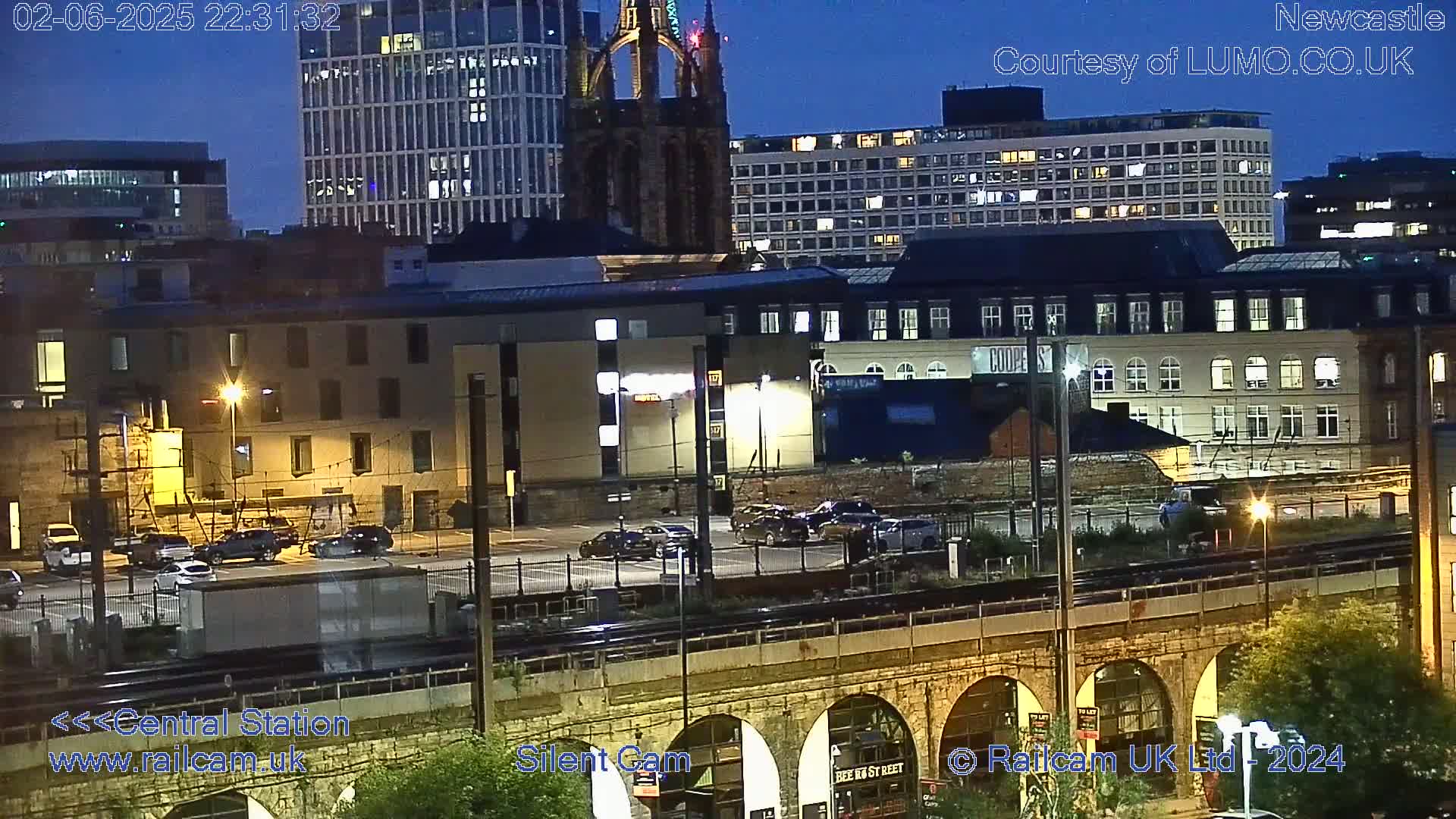 A nighttime view of a city with several buildings illuminated, a parking lot with cars, and a stone arch bridge over train tracks.