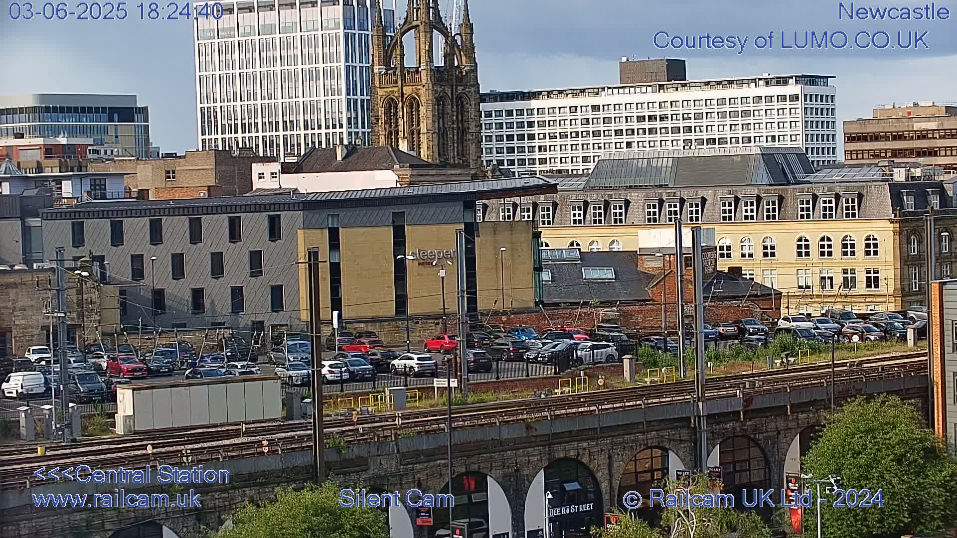 A view of a city with several buildings, including a large church and modern high-rises, a parking lot filled with cars, and train tracks, under a mostly clear sky.