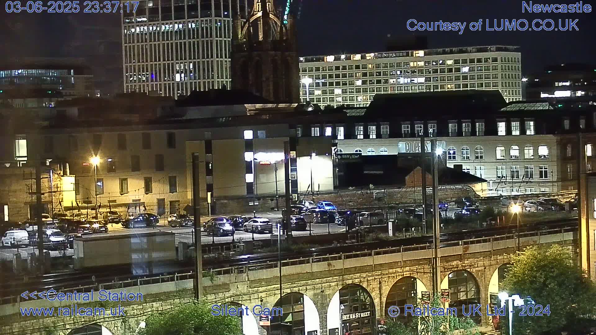 A nighttime view of a city's cityscape, including buildings, a parking lot, train tracks, and a stone arch bridge.