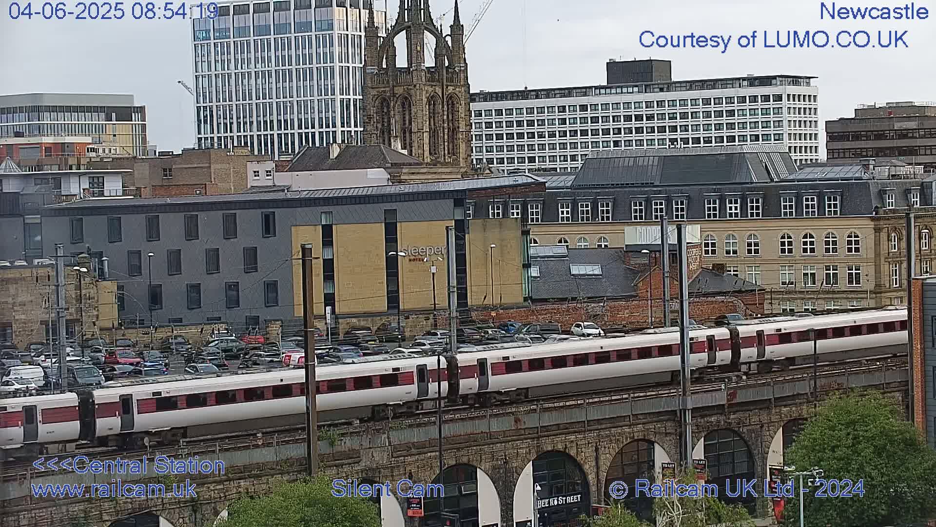 A long, white and maroon train travels across a stone bridge over a city with overcast skies.
