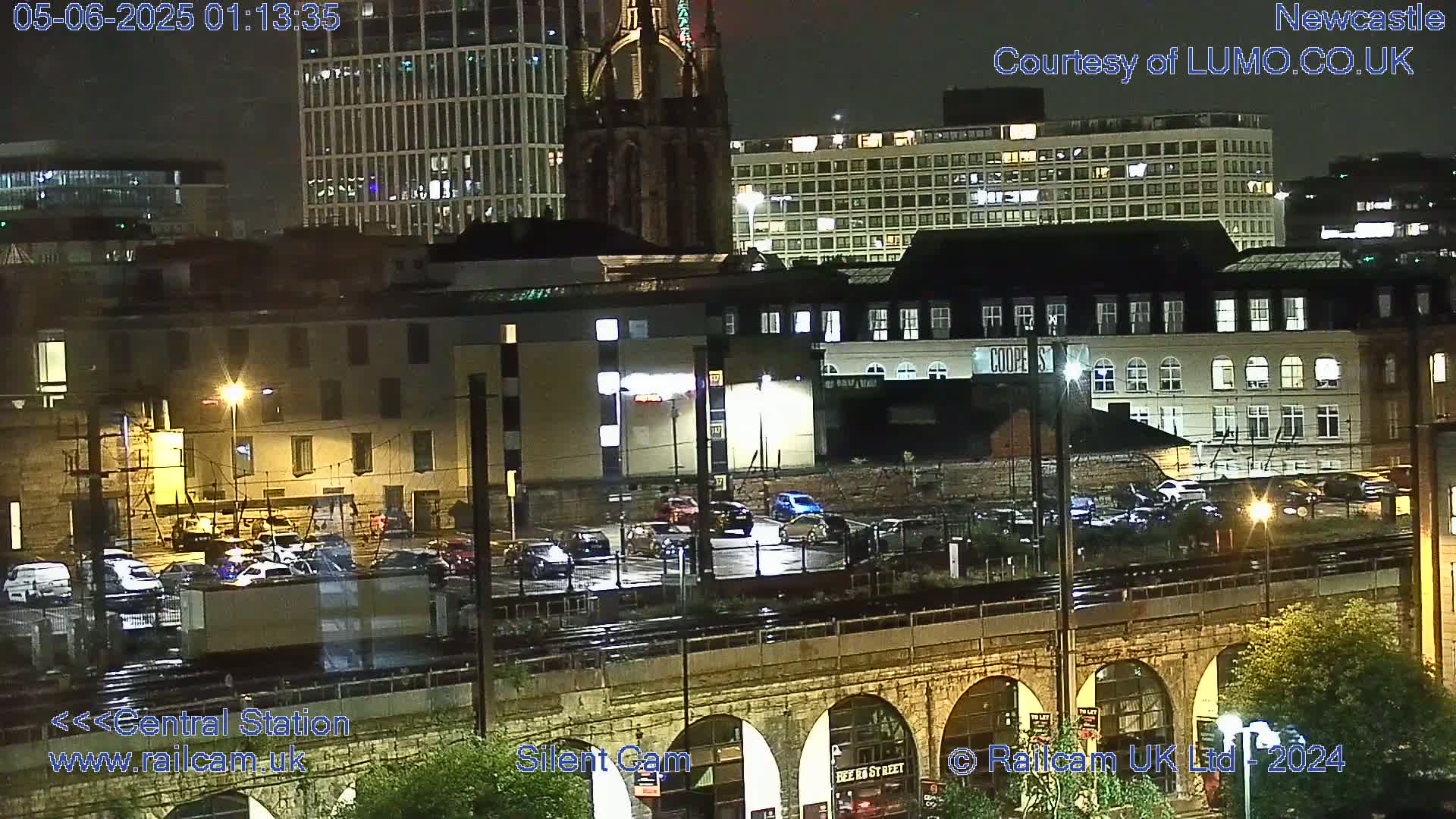 A nighttime view of a city’s railway station area with parked cars, buildings, and a stone arch bridge.