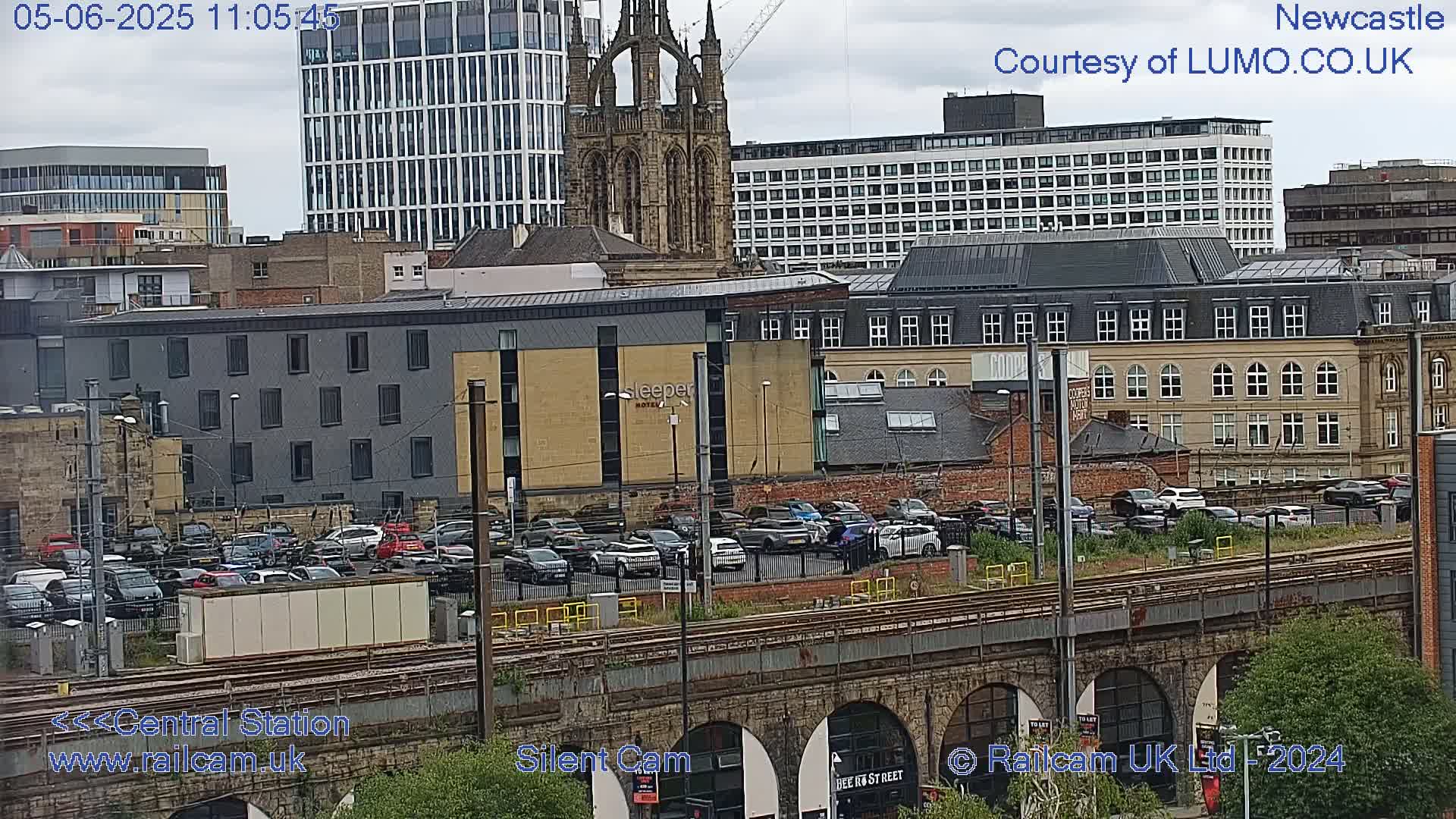 A city scene on an overcast day shows a parking lot full of cars next to train tracks, with various buildings including a tall modern structure and an older Gothic-style church in the background.