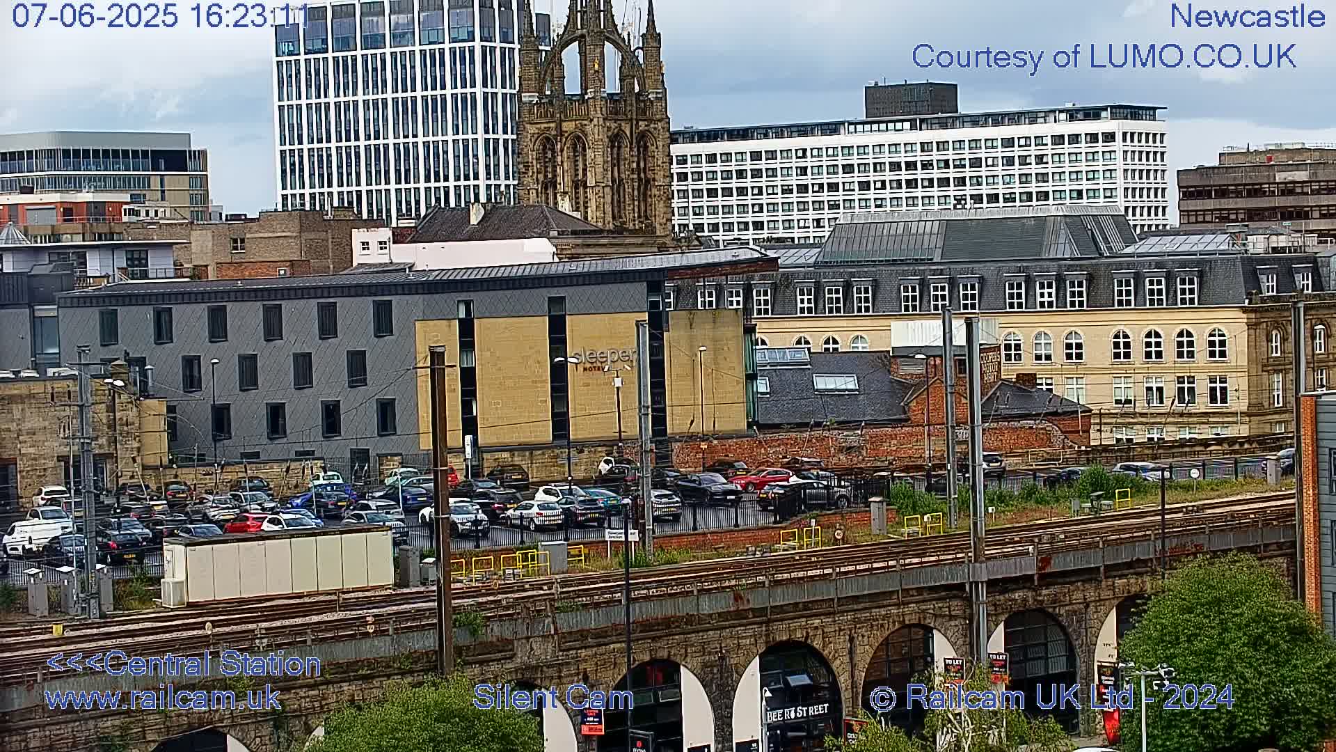 A city scene with overcast skies shows a mix of modern and older buildings, a parking lot full of cars, and train tracks running beneath a stone viaduct.