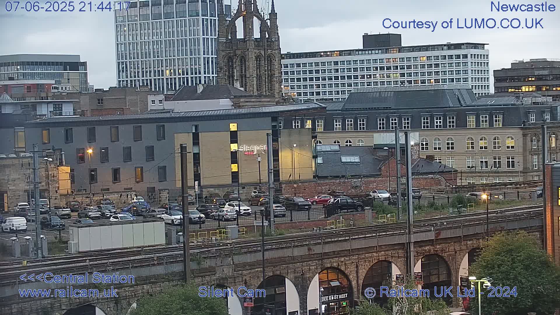 A nighttime view of a city with numerous buildings, including a hotel, a church, and high-rises, alongside train tracks and parked cars under a cloudy sky.