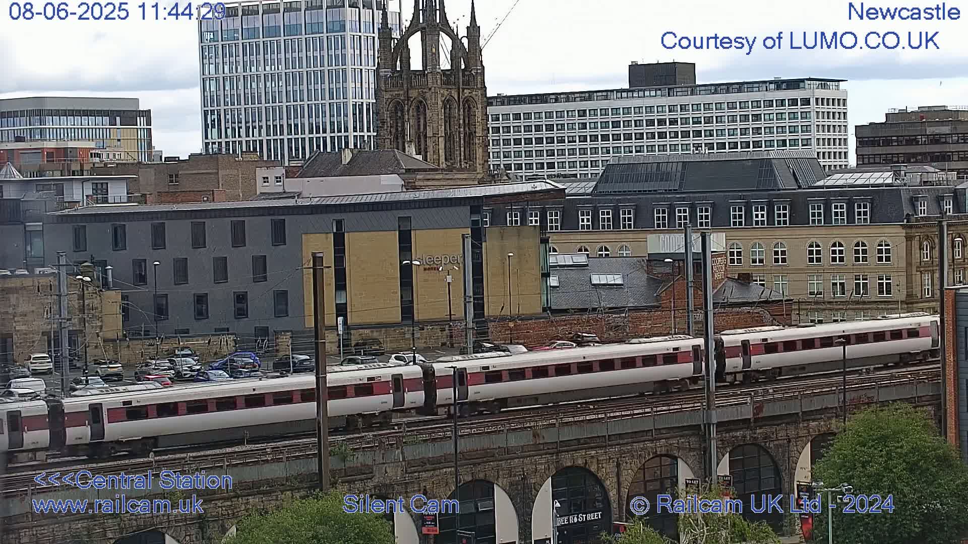 A long passenger train travels across a bridge over a city, under an overcast sky.