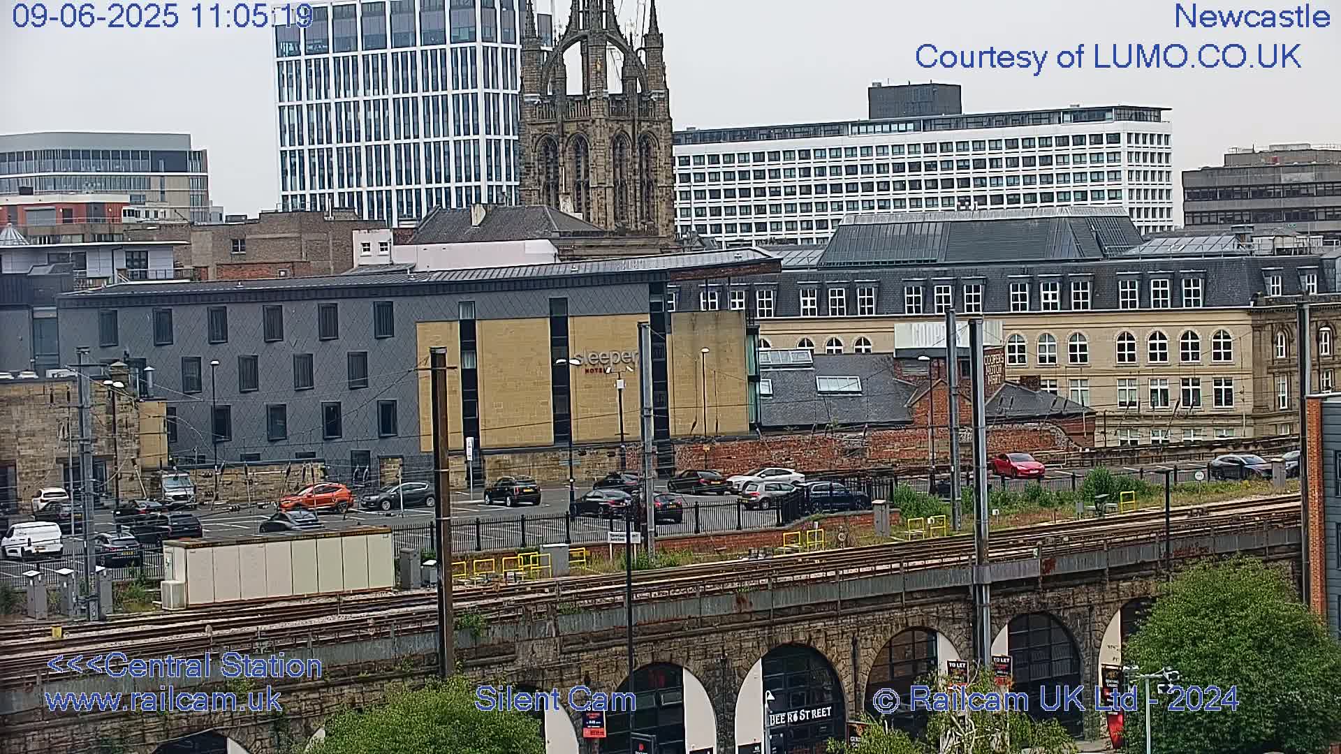 A city scene with overcast skies shows various buildings, including a church and modern high-rises, a parking lot with cars, and train tracks below a stone bridge.
