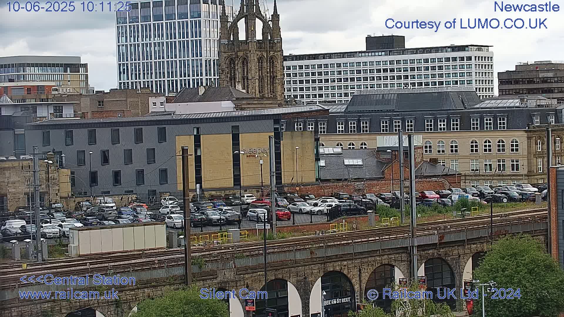 A city scene viewed from above, showing a mix of modern and older buildings, a large parking lot full of cars, and train tracks under a stone bridge, under mostly cloudy skies.