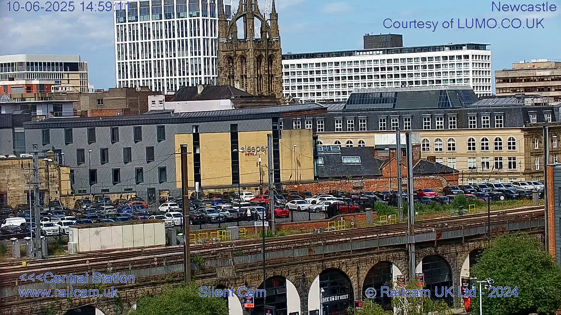 A city view on a partly sunny day shows a large parking lot filled with cars next to a railway bridge and various buildings, including a hotel and a cathedral.