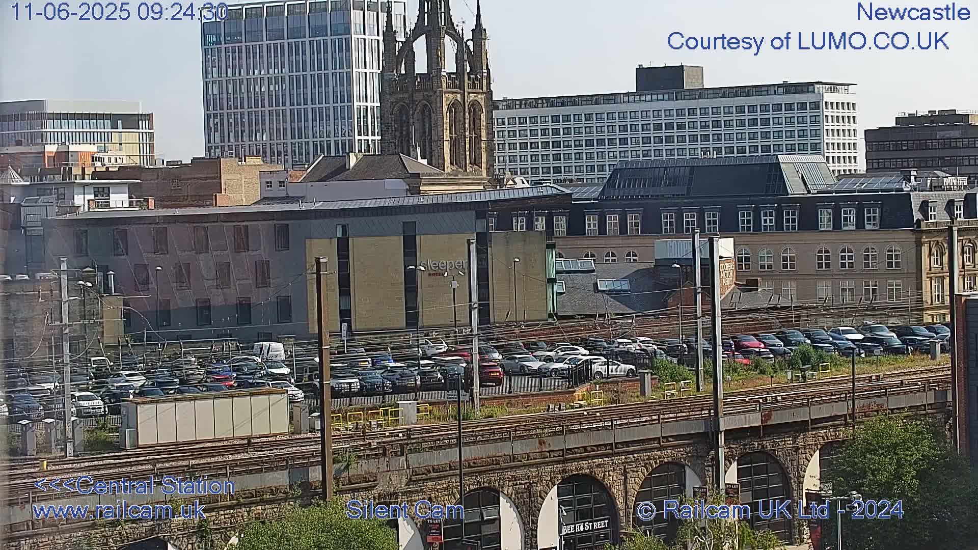 A city view on a sunny day shows a train station with a large parking lot filled with cars, various buildings, and a tall church steeple in the background.