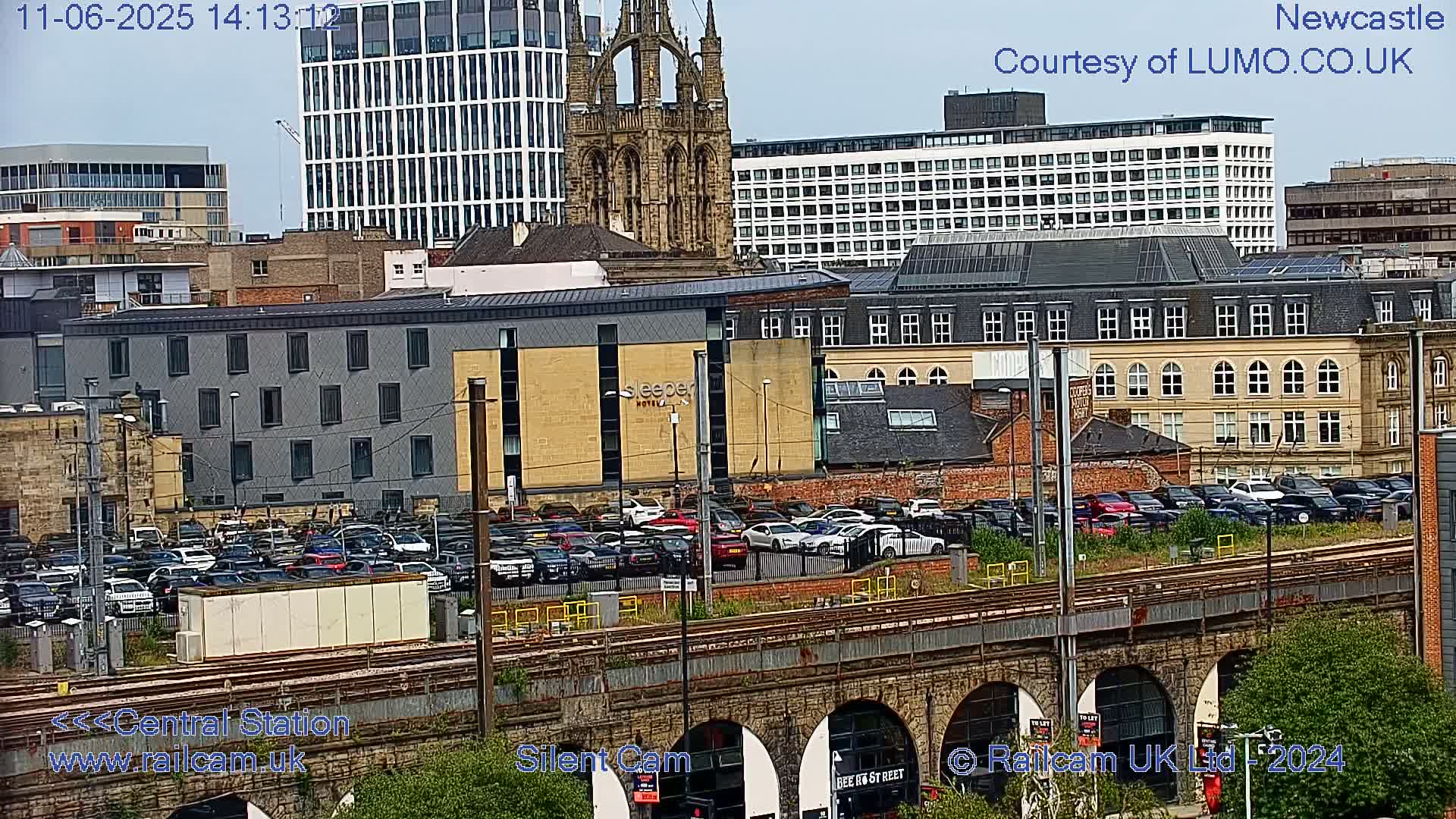A city scene on a partly sunny day shows a railway bridge over a parking lot filled with cars, numerous buildings, and a large church in the background.