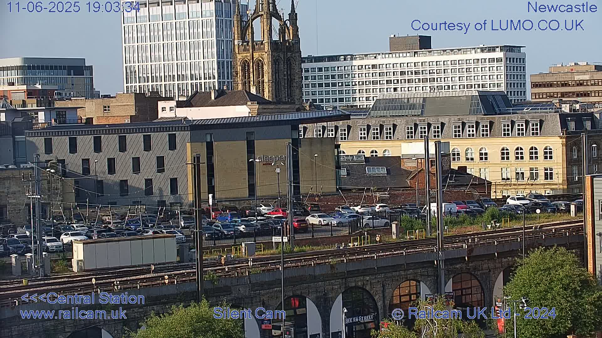 A city view at dusk showing a parking lot, train tracks, various buildings including a tall church, and some greenery.