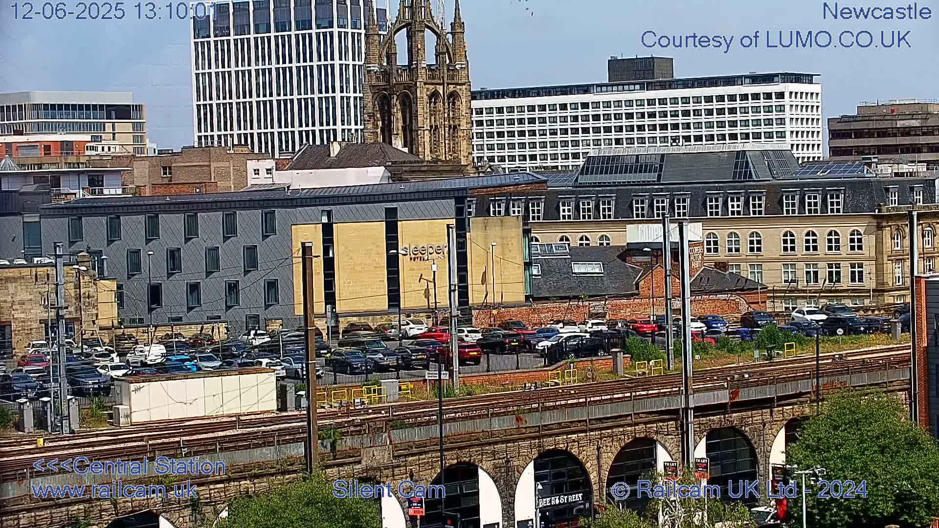 A city scene on a sunny day shows a parking lot full of cars next to train tracks that run under a series of stone arches, with various buildings including a hotel and a church visible in the background.