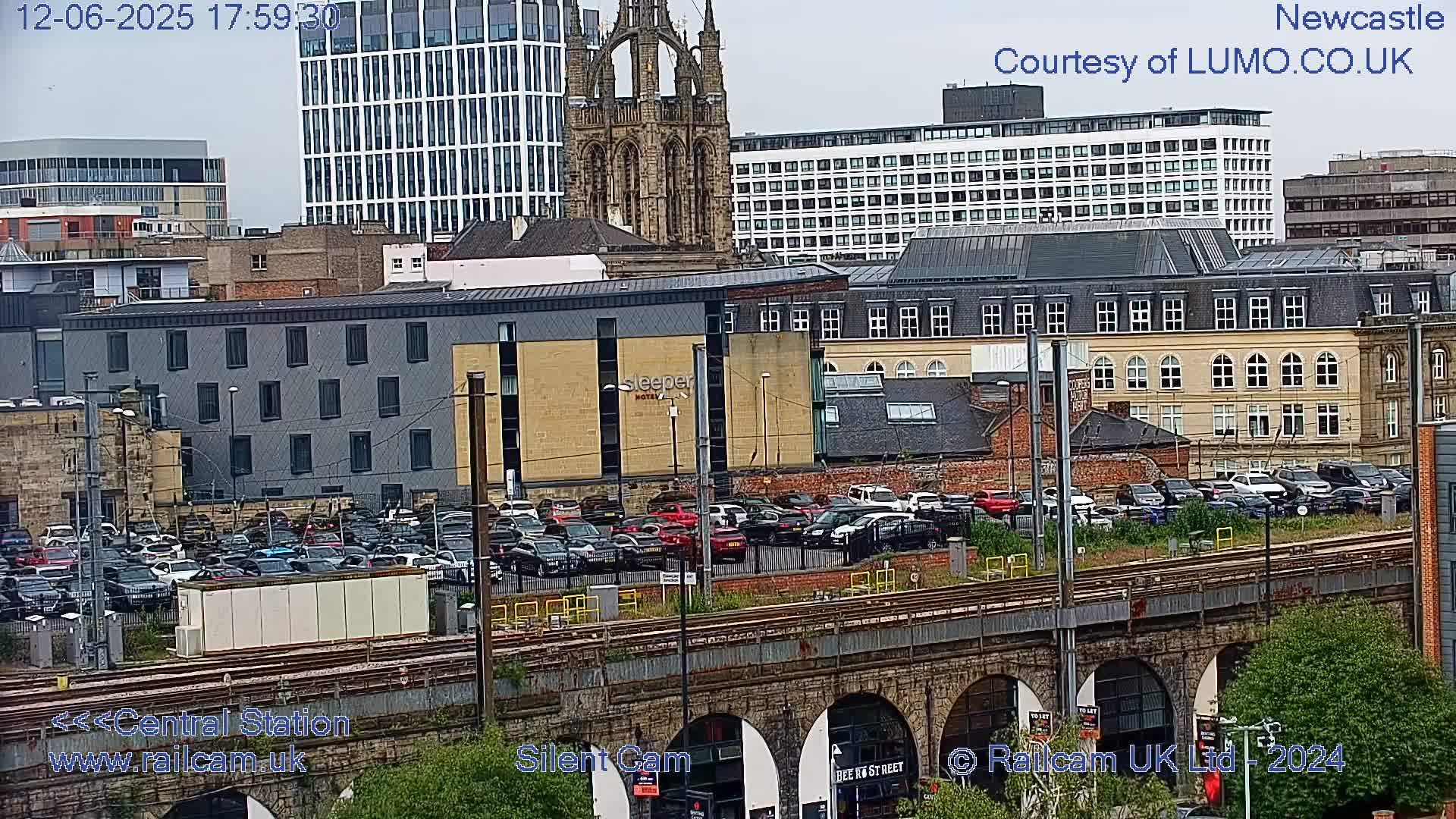 A city view with overcast skies shows a parking lot full of cars next to a train track that runs under a stone archway bridge, all in front of a variety of buildings including a hotel and a church.