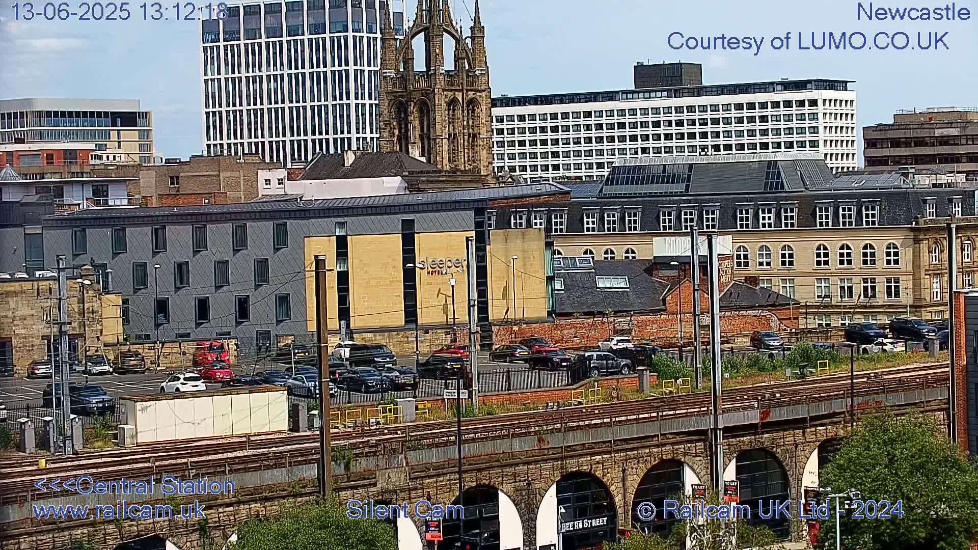 A city view on a sunny day shows a mix of modern and older buildings, a parking lot filled with cars, and railway tracks and bridges below.